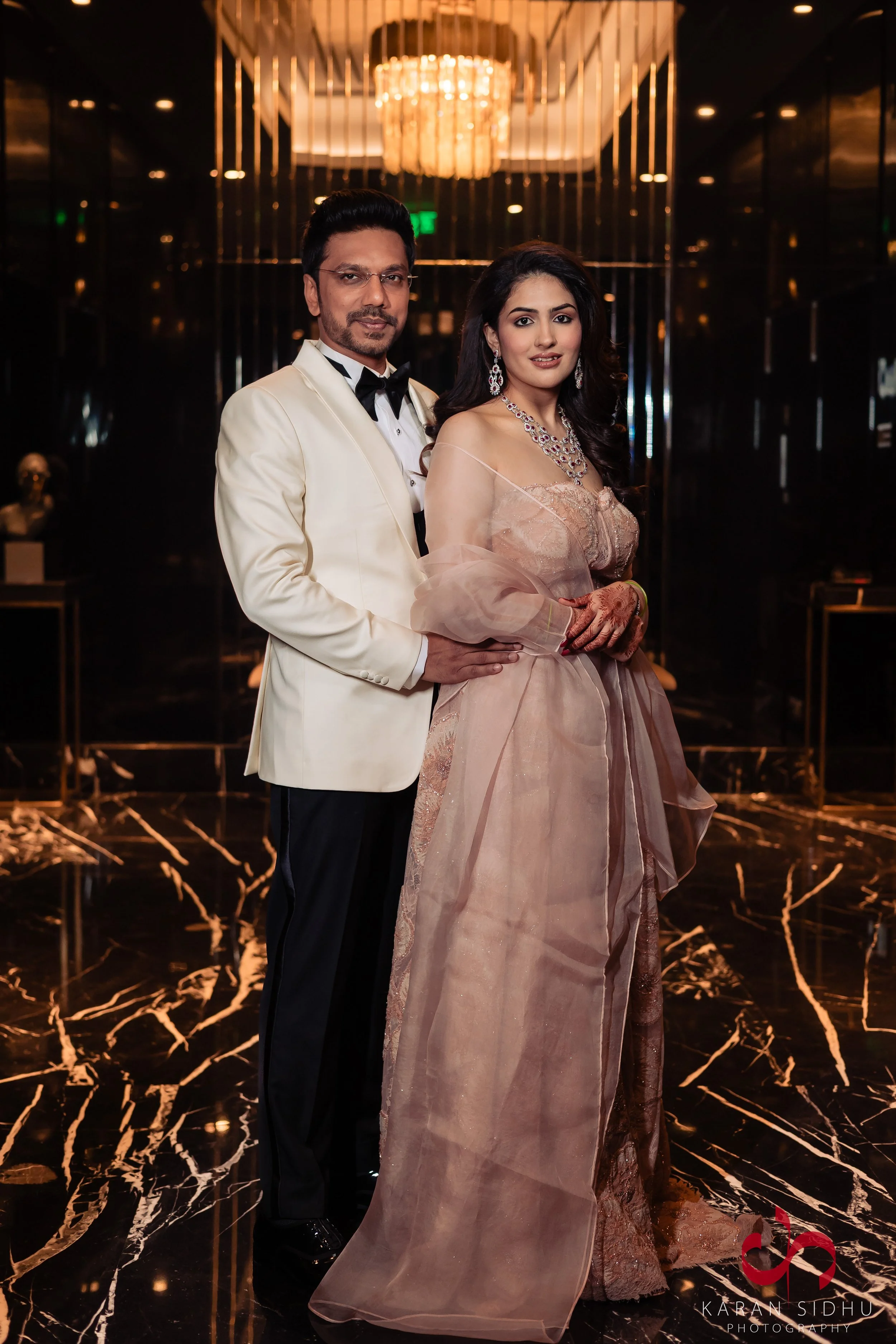 A well-dressed couple posing for a photograph in an elegant, dimly lit venue with a chandelier overhead and black and gold decorative elements in the background.
