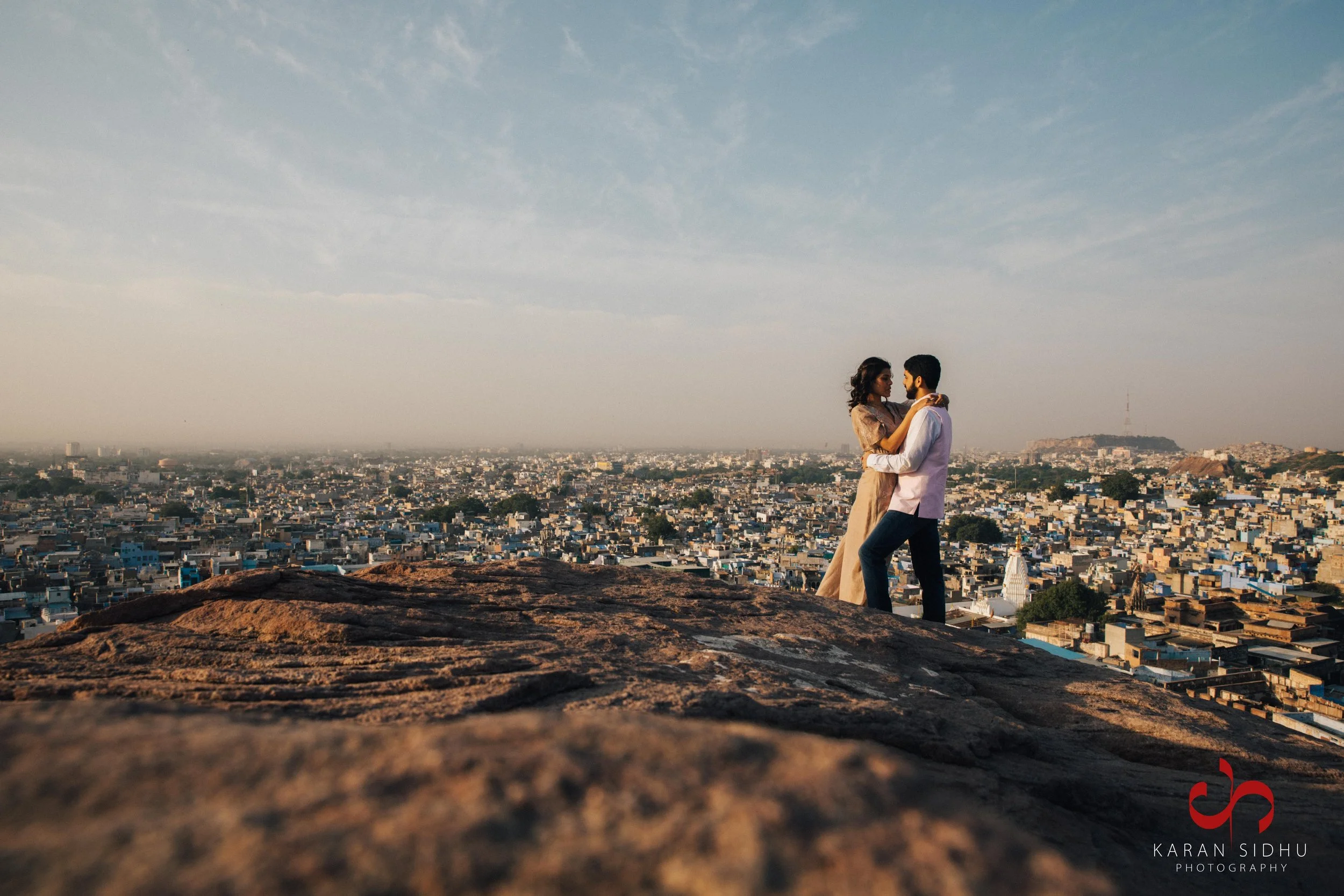 A couple stands on a hilltop overlooking a cityscape at sunset, holding each other in a dance-like pose.