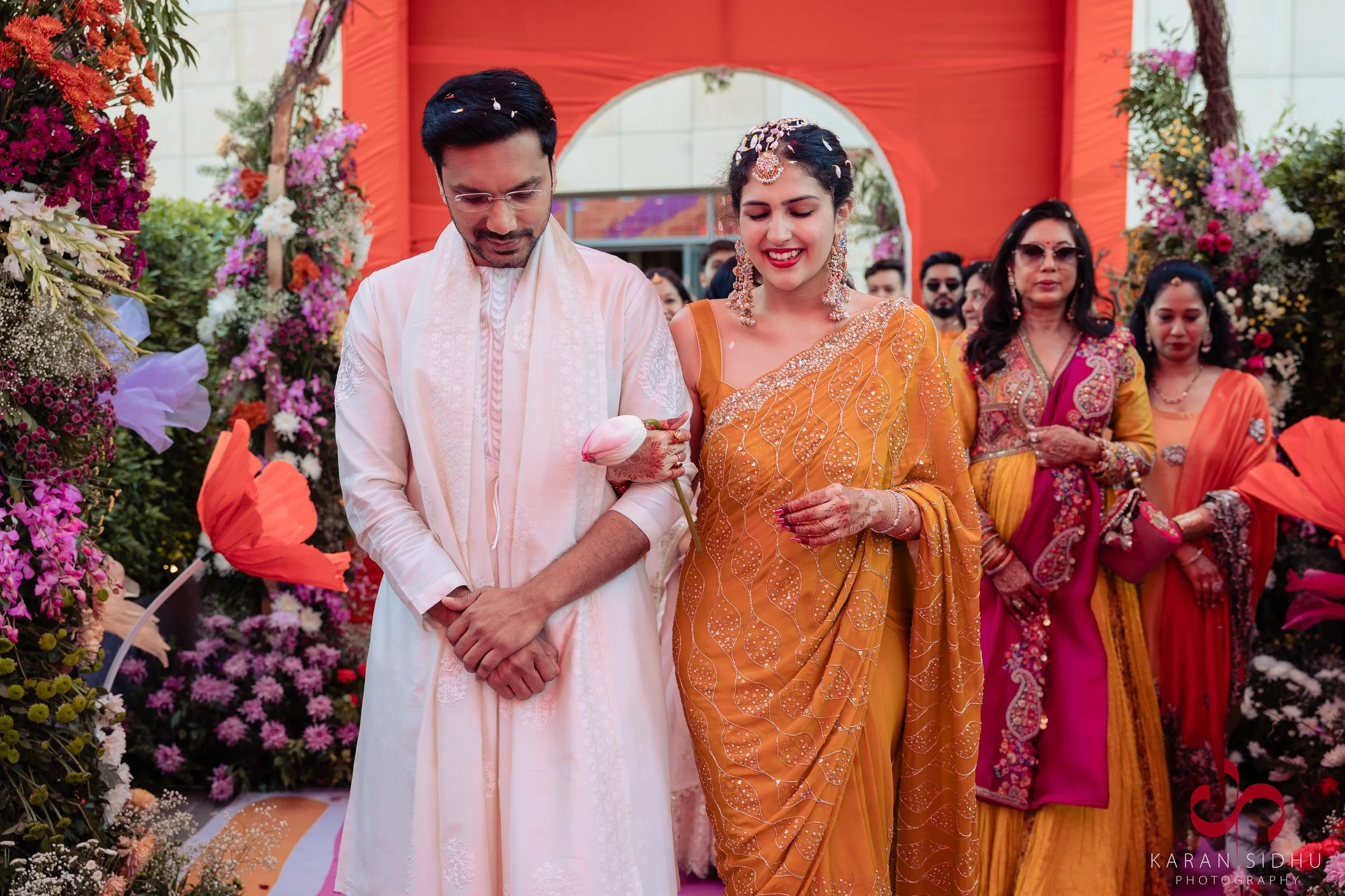 Indian man and woman in traditional wedding attire walk together during a wedding celebration, surrounded by colorful flowers and decorated arch.