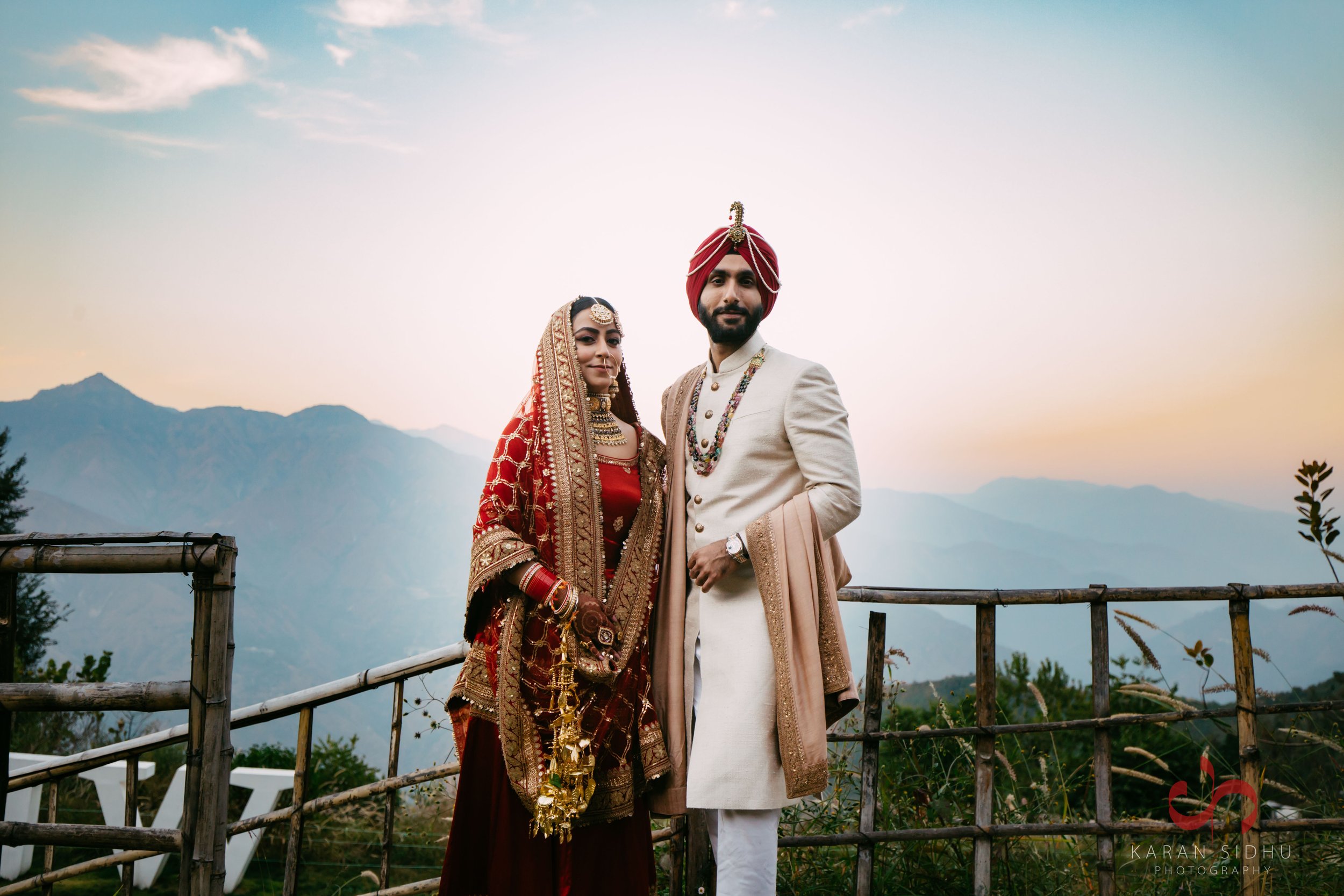 A bride and groom in traditional Indian wedding attire standing outdoors with mountains in the background at sunset.