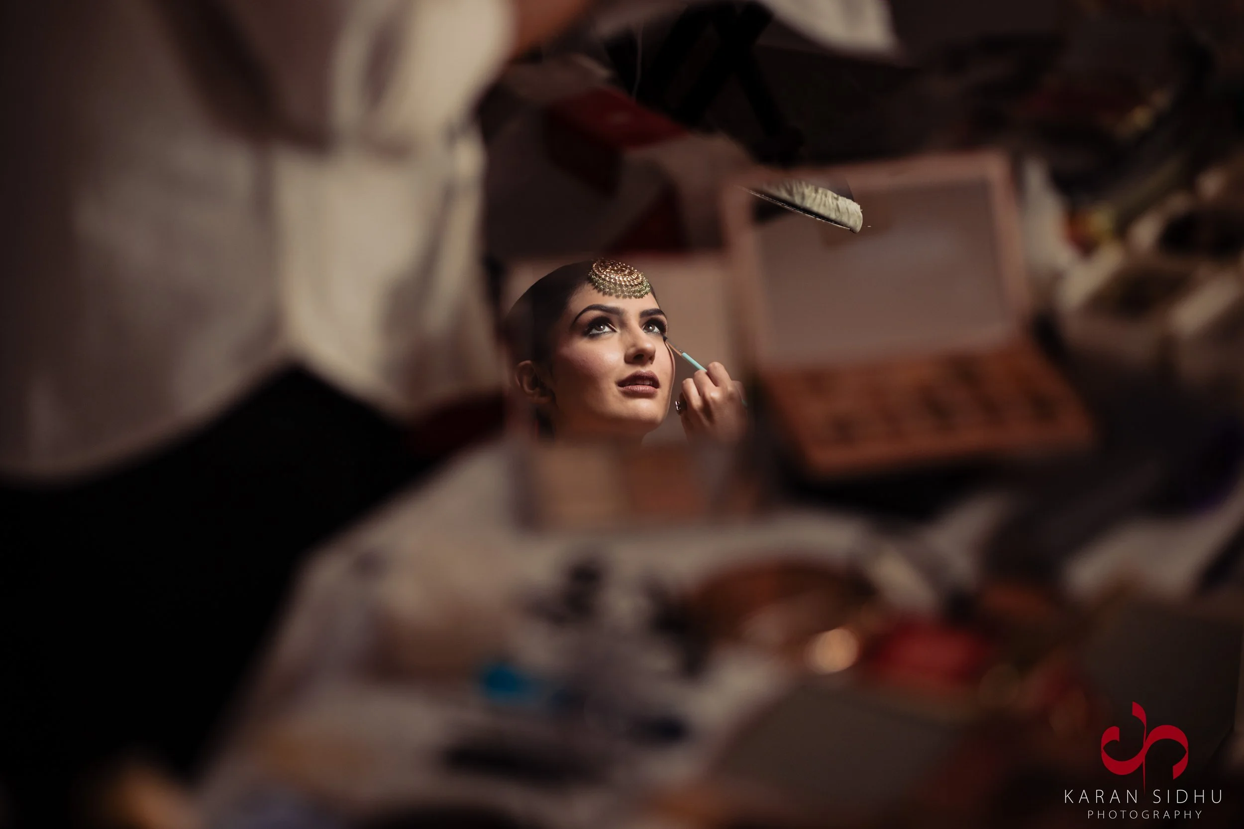 A woman getting her makeup done, seen through a cluttered vanity mirror filled with makeup products and brushes, wearing traditional jewelry with a focused expression.