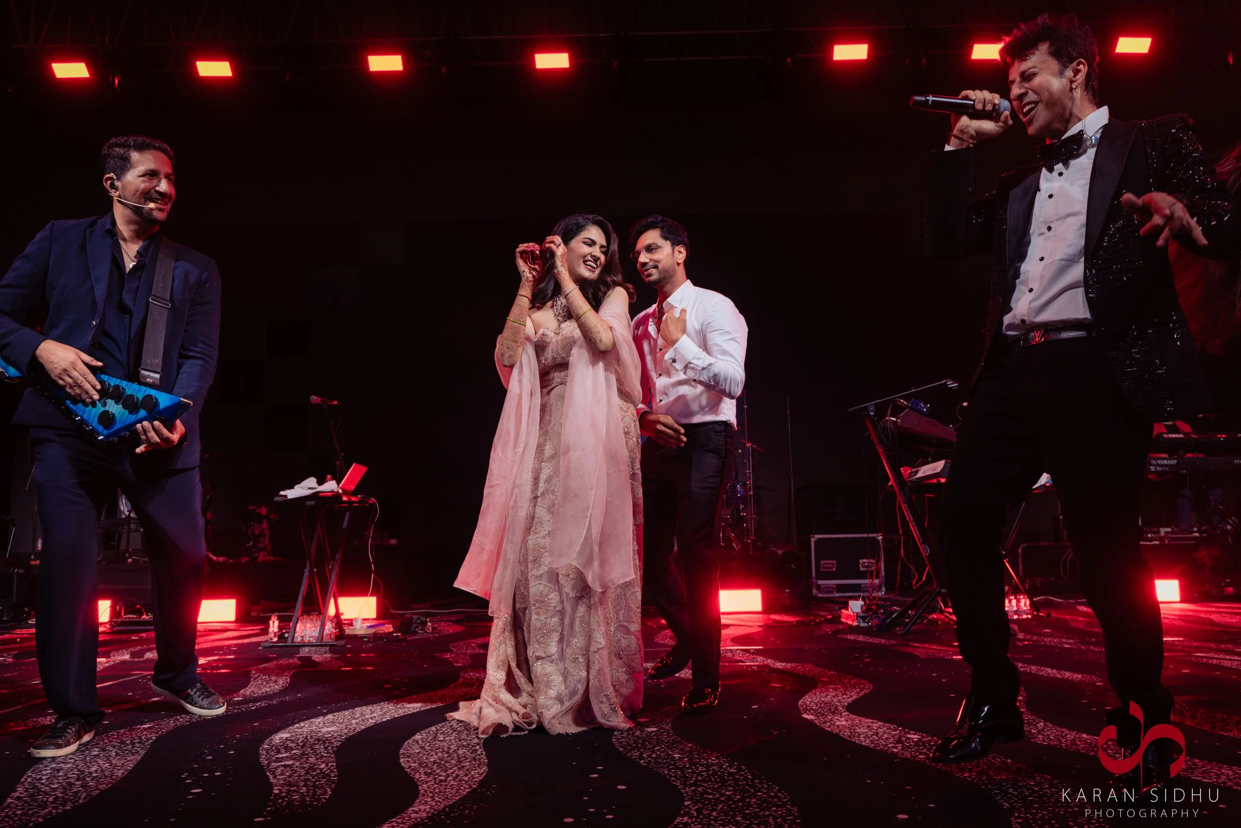 Group of people performing on stage with musical instruments, including a singer with a microphone, a man playing a melodica, and two other men, one in a tuxedo, in front of red stage lights.