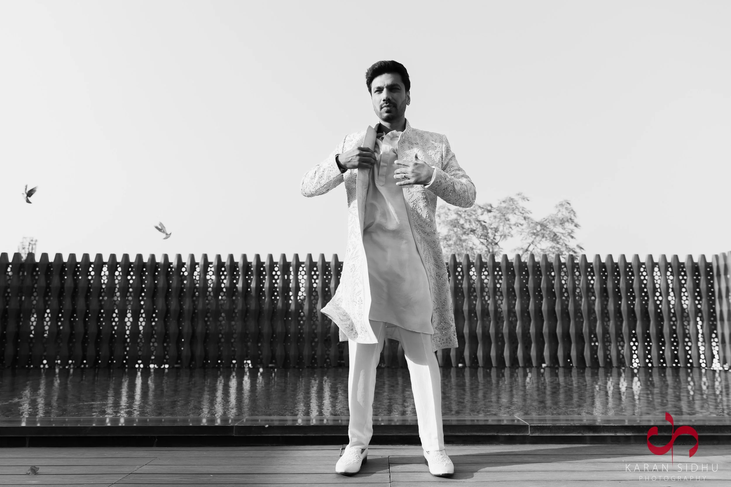 A man in traditional Indian attire standing outdoors with birds flying in the sky behind him.