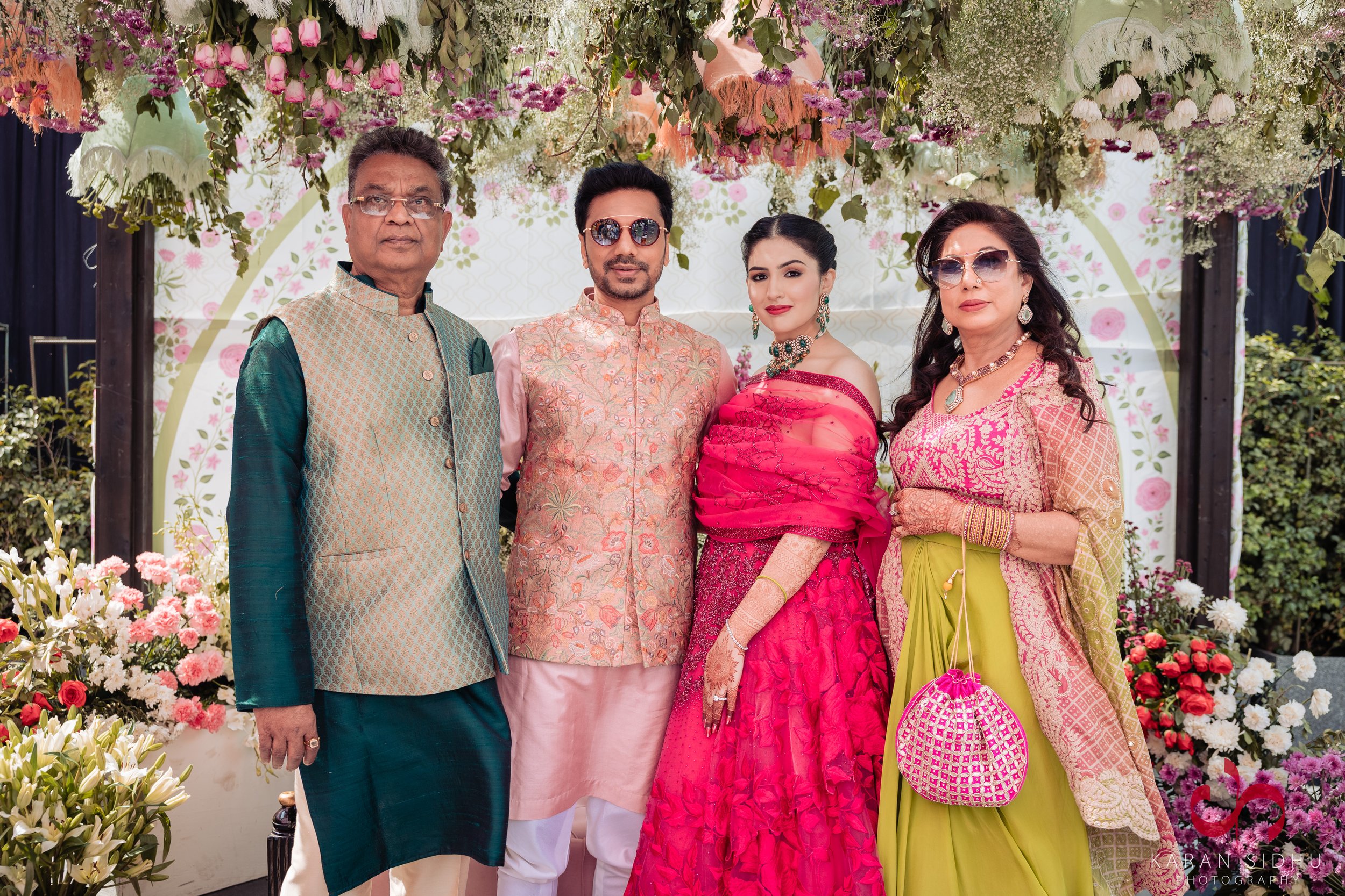 A family of five dressed in traditional Indian attire, standing in front of a floral decorated backdrop at a celebration or wedding event.
