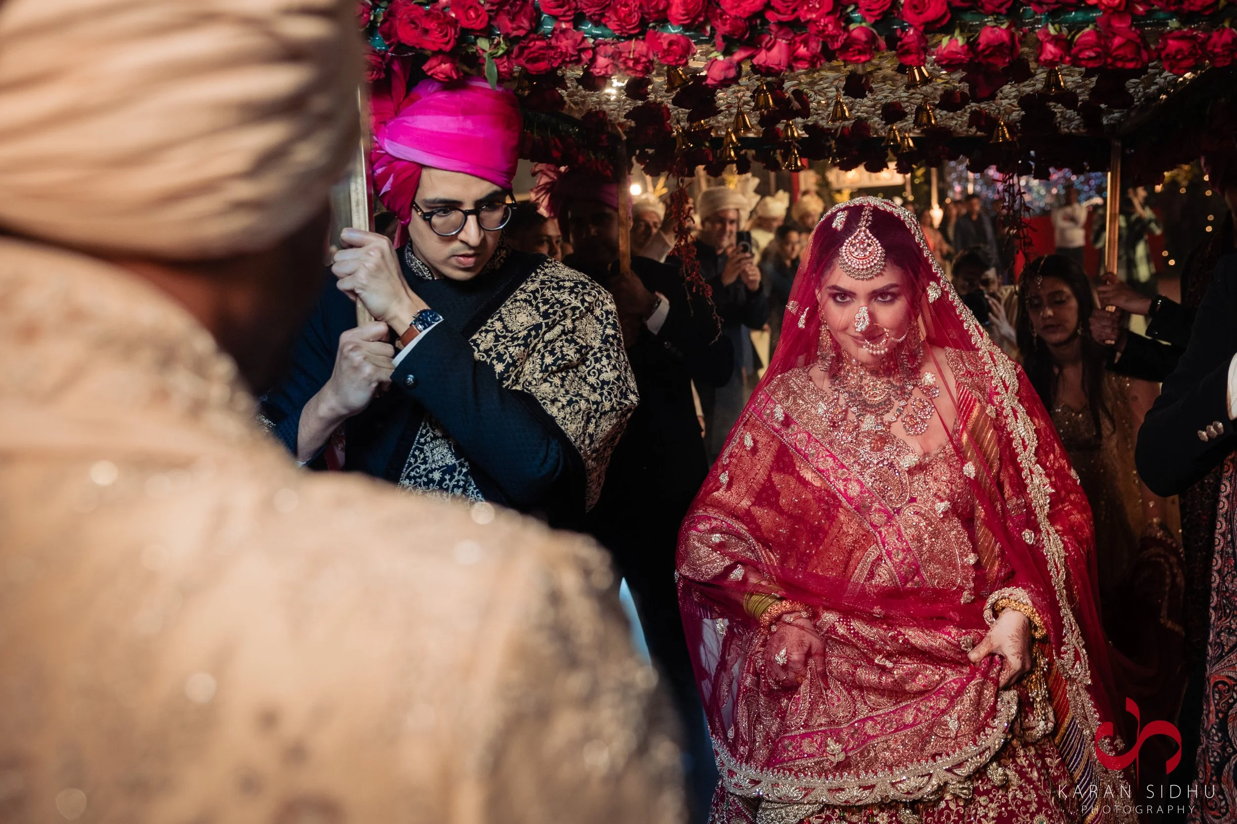 Indian bride in red and gold traditional wedding attire, wearing intricate jewelry and a red veil, under a floral decorated canopy. Groom and guests dressed in traditional attire surround her during the wedding ceremony.