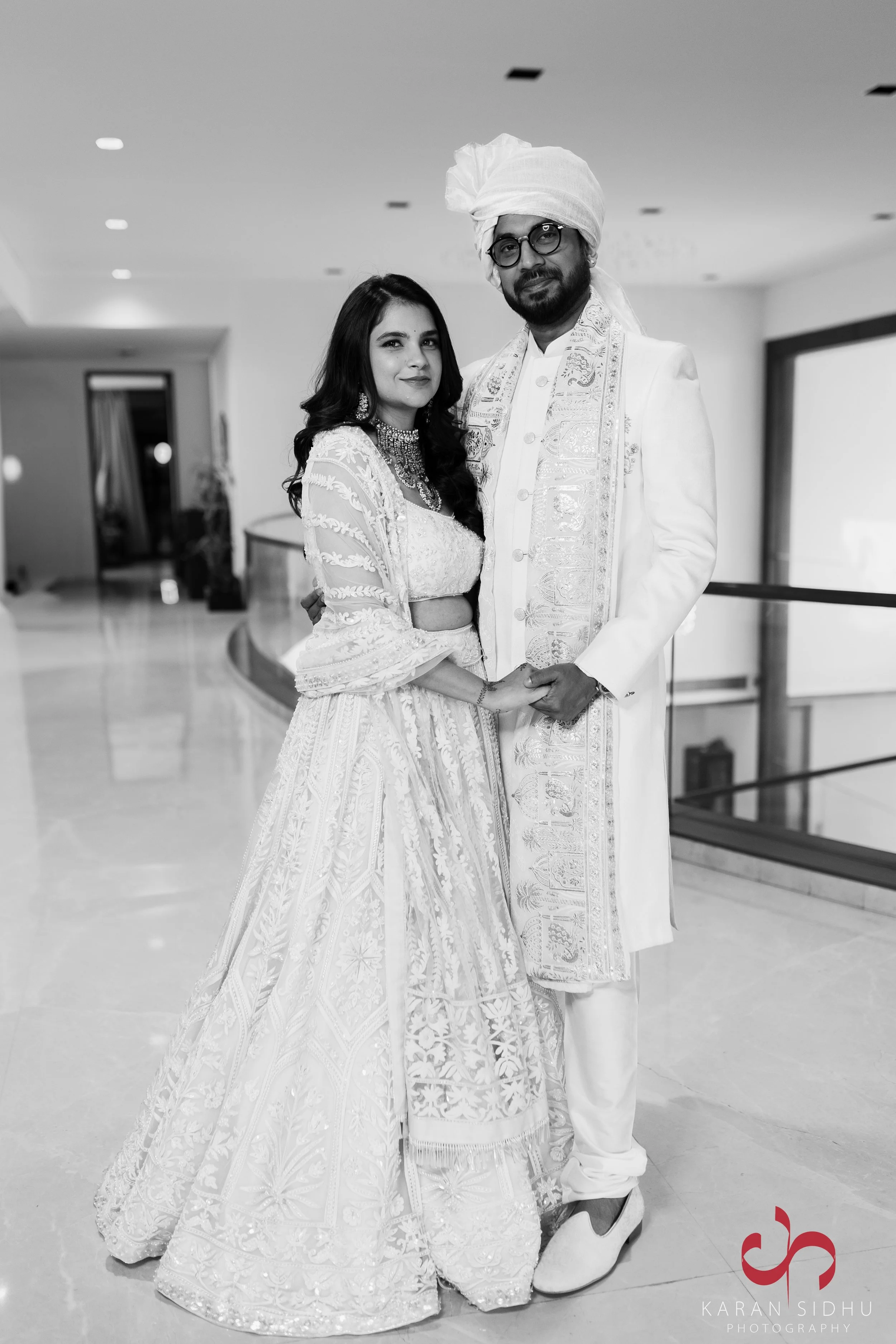 Black and white photo of a couple in traditional Indian wedding attire, standing indoors. The woman wears a heavily embroidered lehenga and jewelry, while the man wears a sherwani and turban. They are holding hands and looking at the camera.