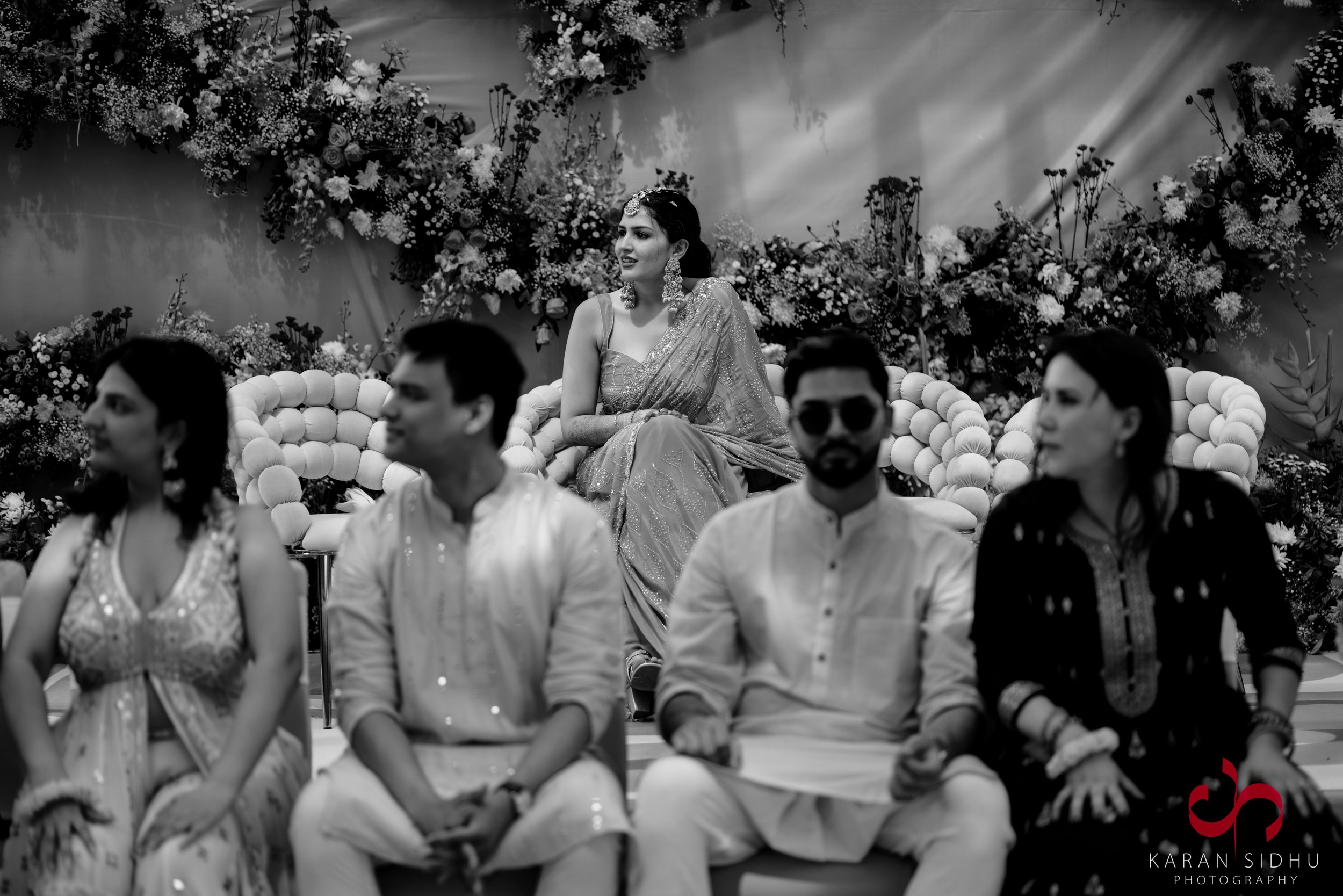 A group of people sitting with a woman in traditional attire sitting on a throne-like chair behind them, with floral decorations on a stage in the background.