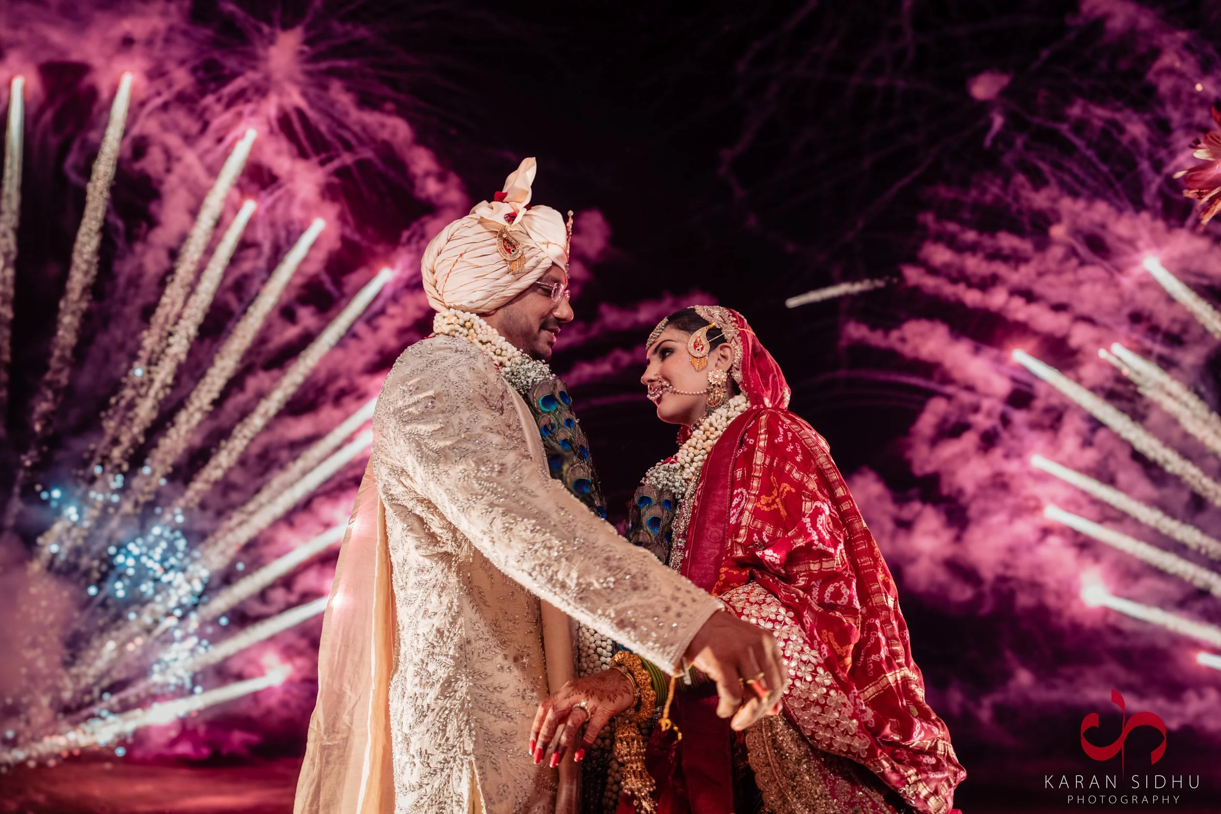 A bride and groom in traditional Indian wedding attire beneath fireworks at night.