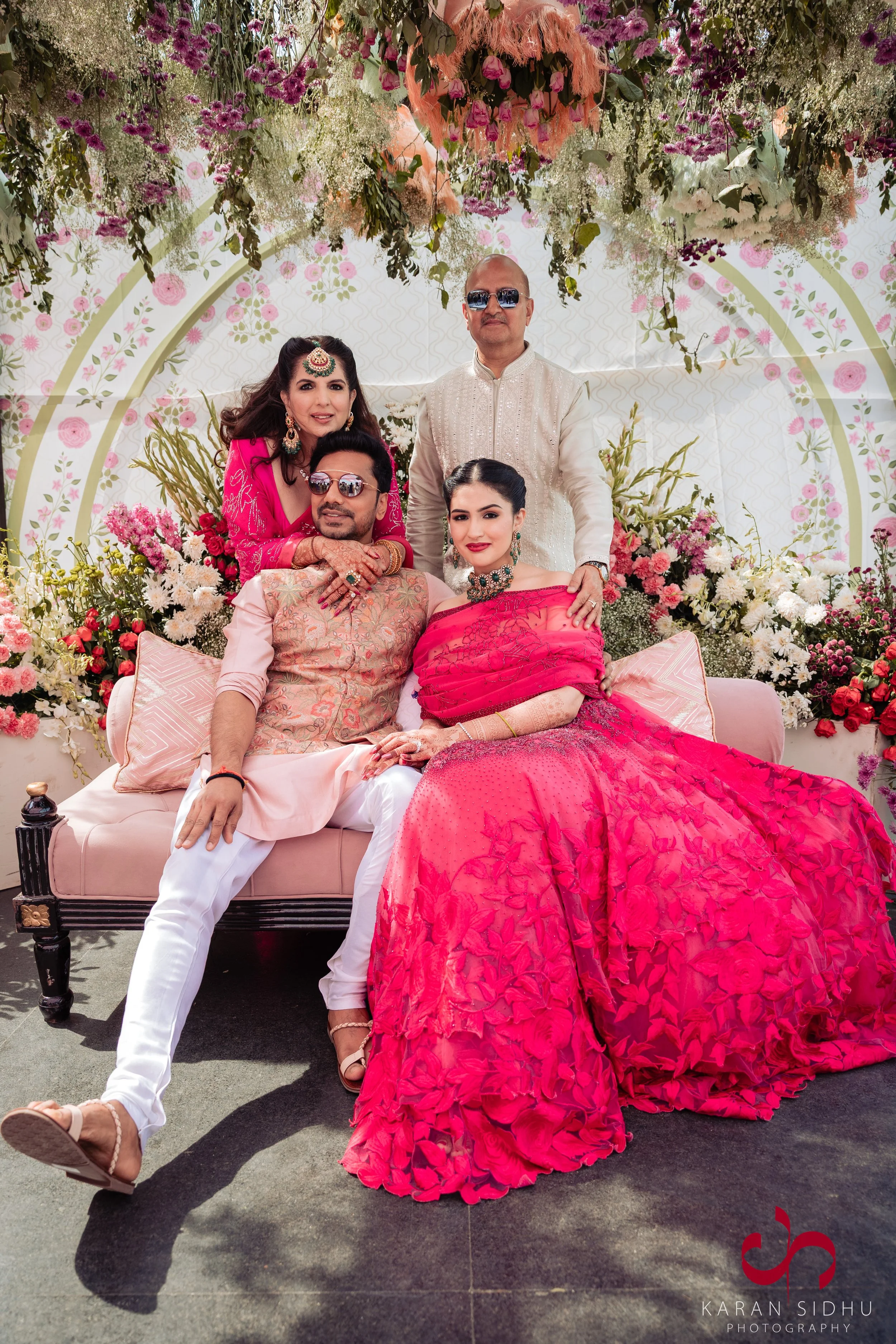 A group of five people dressed in traditional Indian attire sitting and standing on a decorated stage with flowers. The women are wearing pink sarees with jewelry, and the men are wearing kurta-style outfits. The background features floral decoration