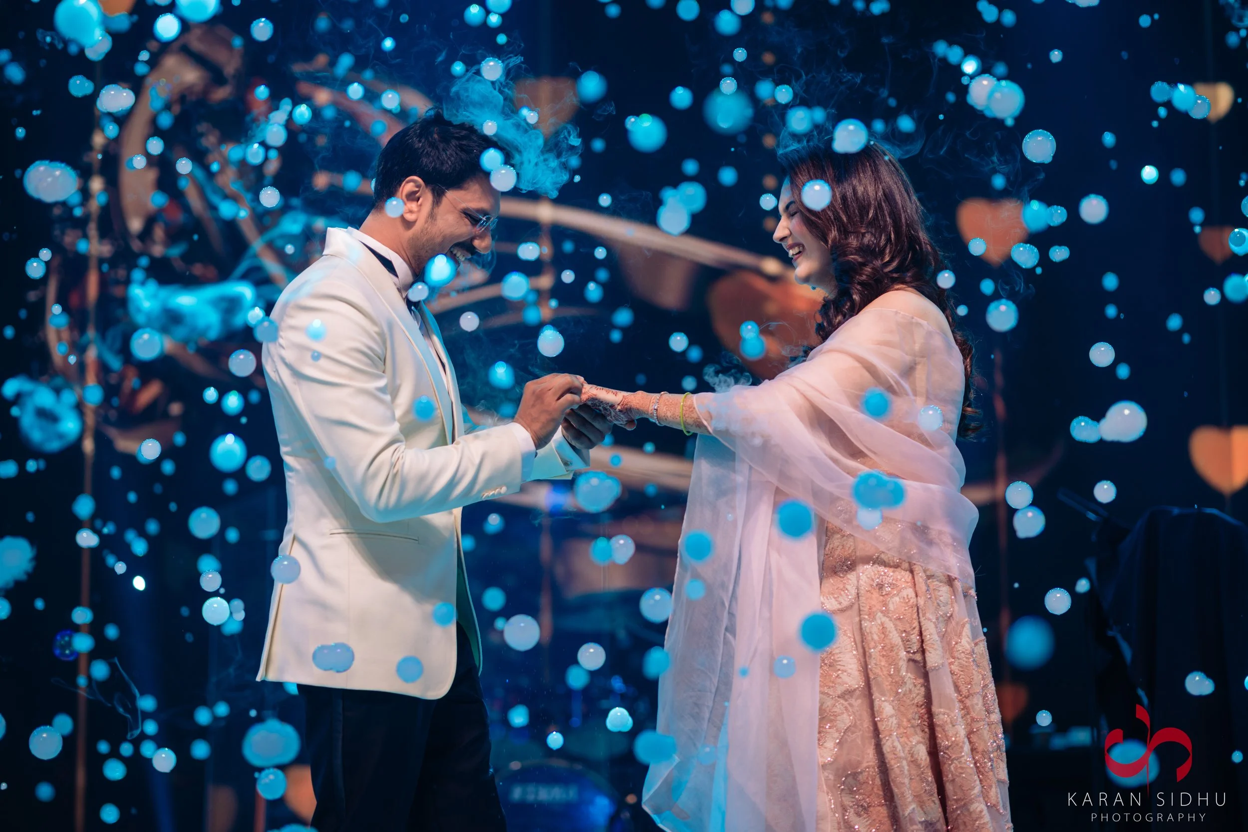 A couple exchanging rings during a wedding ceremony, with blue confetti falling around them and a dark background.