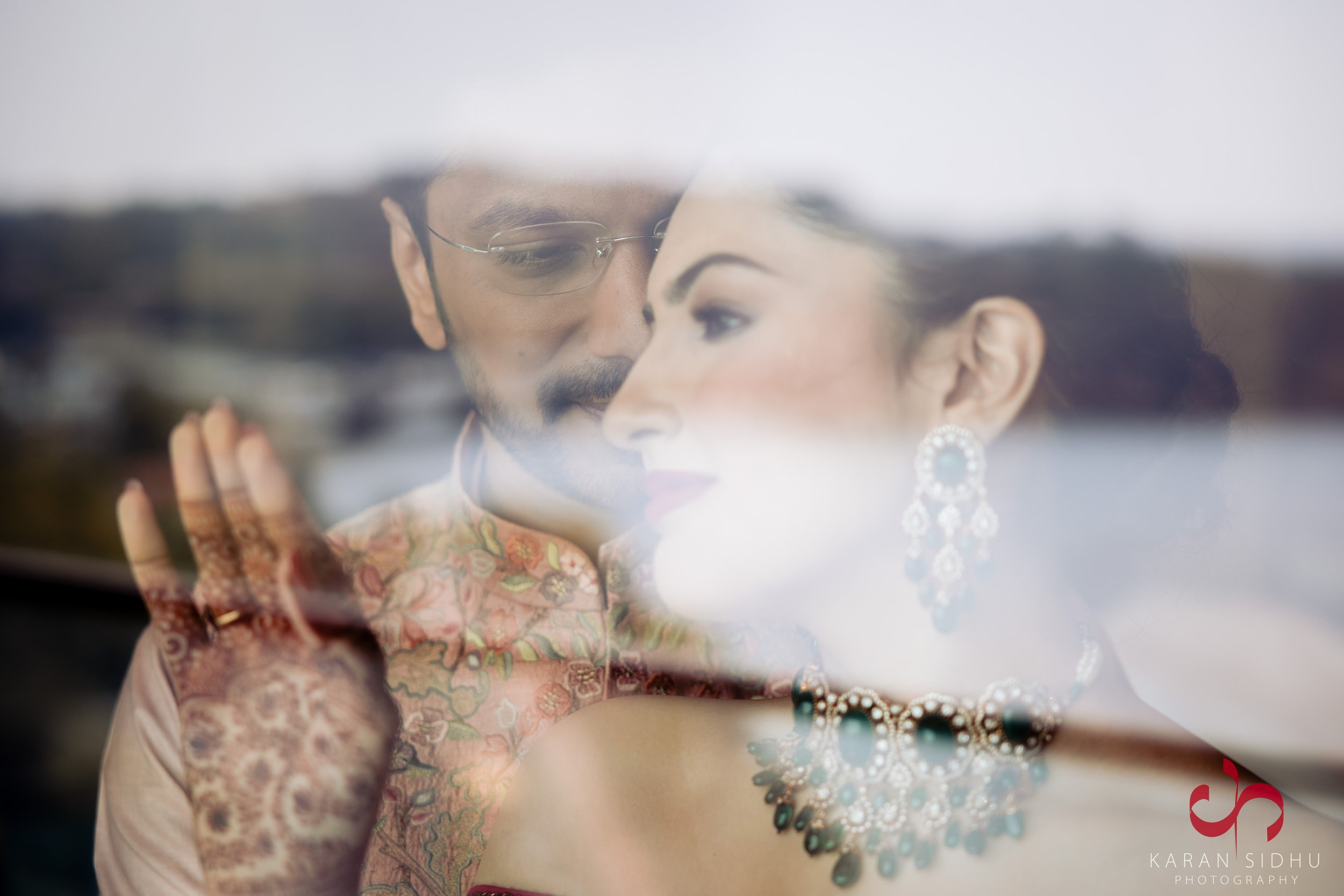 A romantic reflection of a couple dressed in traditional Indian attire, with the woman wearing earrings and a necklace, and the man wearing glasses and a sherwani, both looking at each other through a glass window.