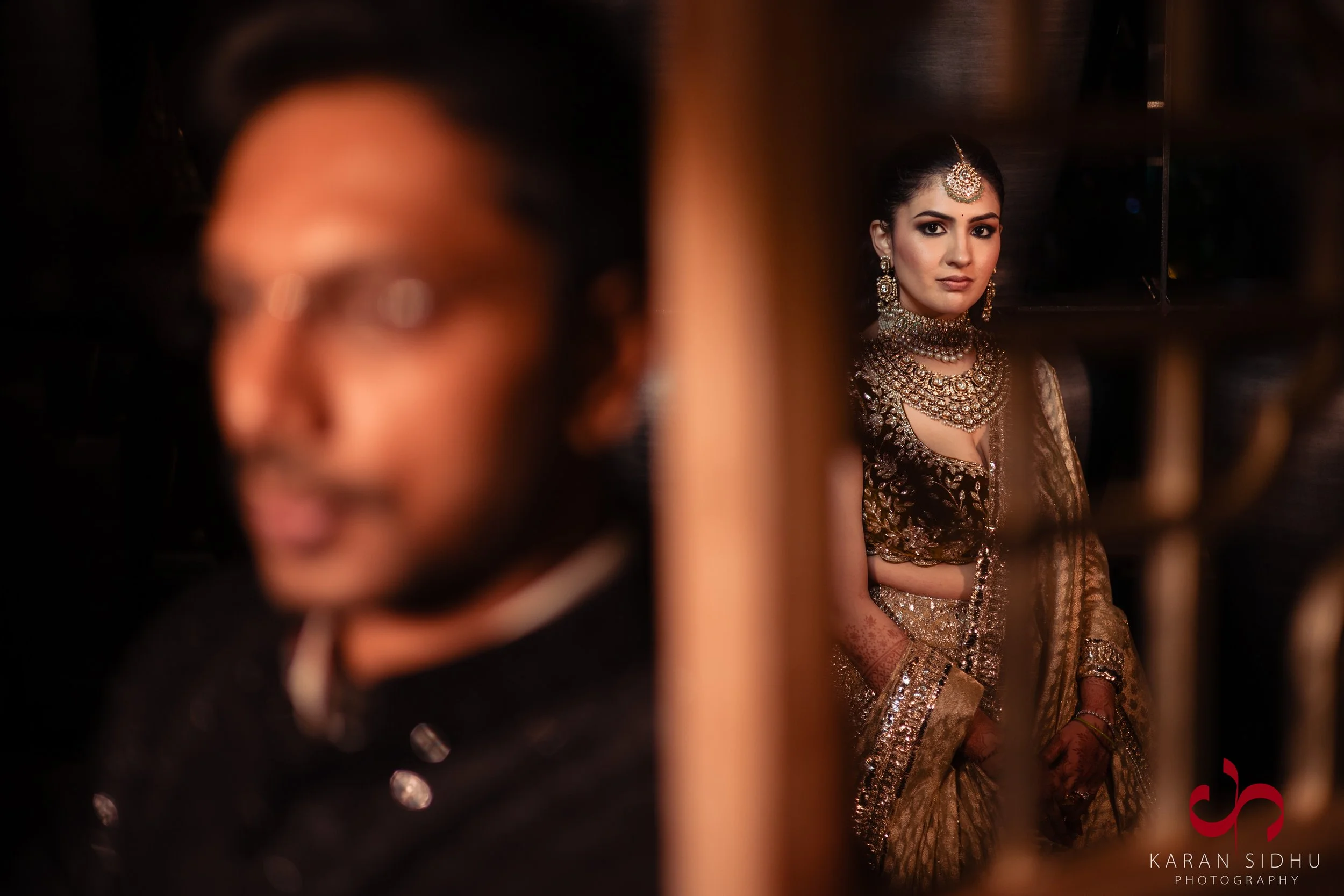 A woman dressed in traditional Indian bridal attire, adorned with elaborate jewelry including a necklace, earrings, and a maang tikka, poses gracefully. A man, blurred in the foreground, appears to be her partner, dressed in formal attire. The woman'