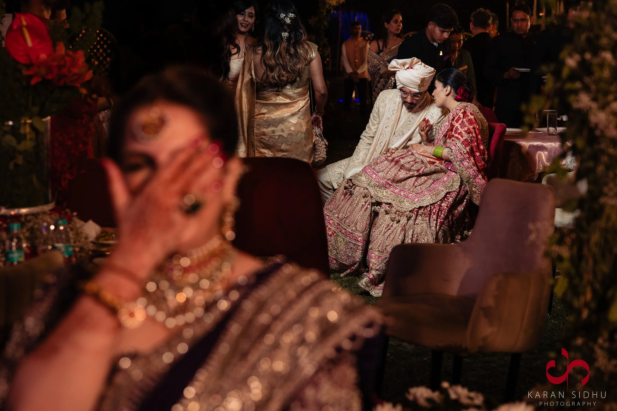 People dressed in traditional Indian attire at a wedding celebration, with some guests smiling and engaging in conversations, and a woman in the foreground covering her face with her hand.