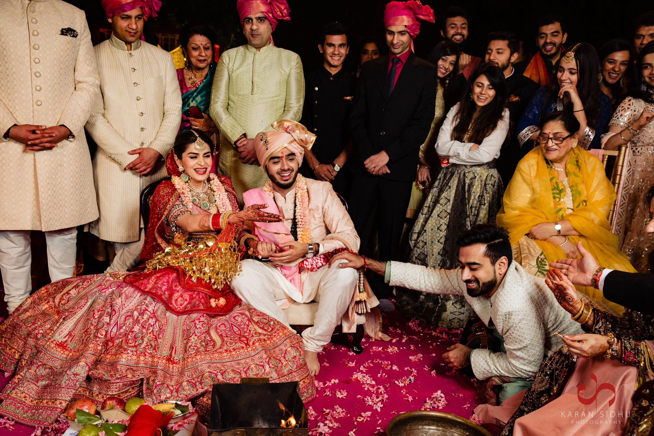 Indian wedding ceremony with the bride and groom sitting on a decorated floor, surrounded by family and friends. The bride wears a red and gold traditional dress with jewelry, and the groom wears a cream-colored outfit with a pink sash and turban. The guests are dressed in traditional attire and are smiling and participating in the celebration.