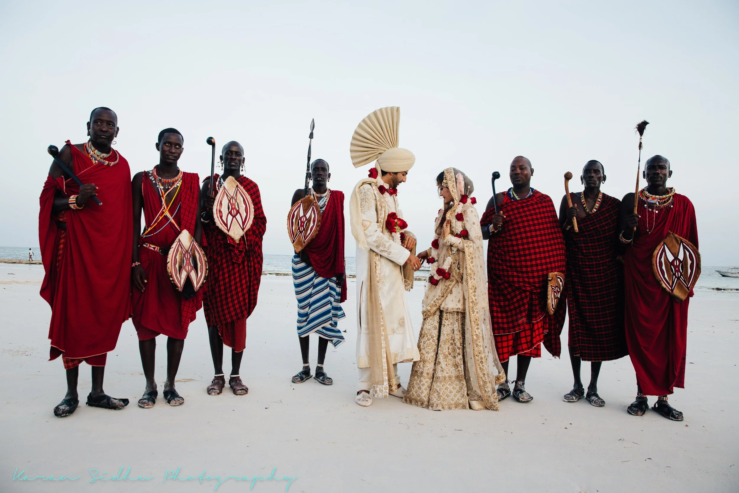 A wedding ceremony on the beach featuring an African bride and groom dressed in traditional attire, surrounded by men in red and black traditional Maasai clothing, holding spears and shields.