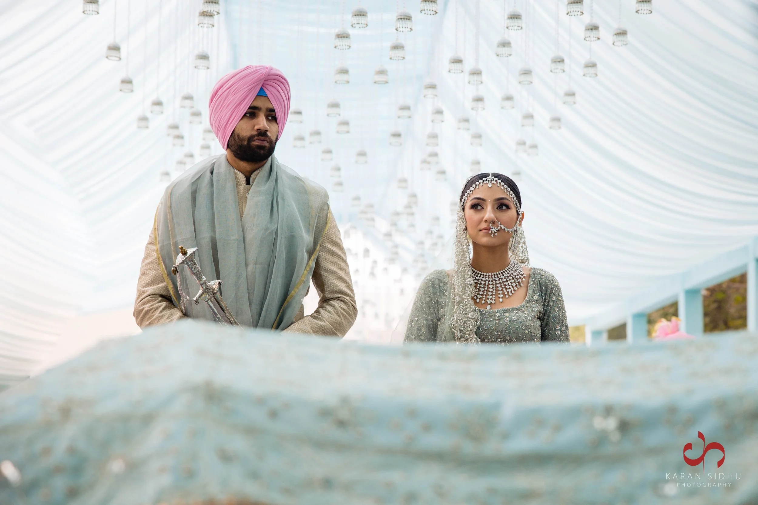 A bride and groom at a wedding ceremony, dressed in traditional Indian attire, standing under a decorated canopy with hanging lanterns.