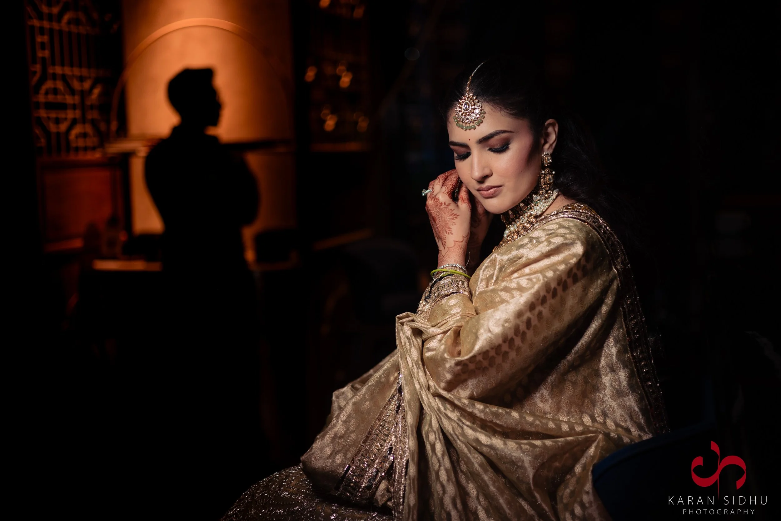 A woman dressed in traditional Indian attire, wearing jewelry, with henna on her hands, sitting with her eyes closed, in a dimly lit setting with a person's silhouette in the background.