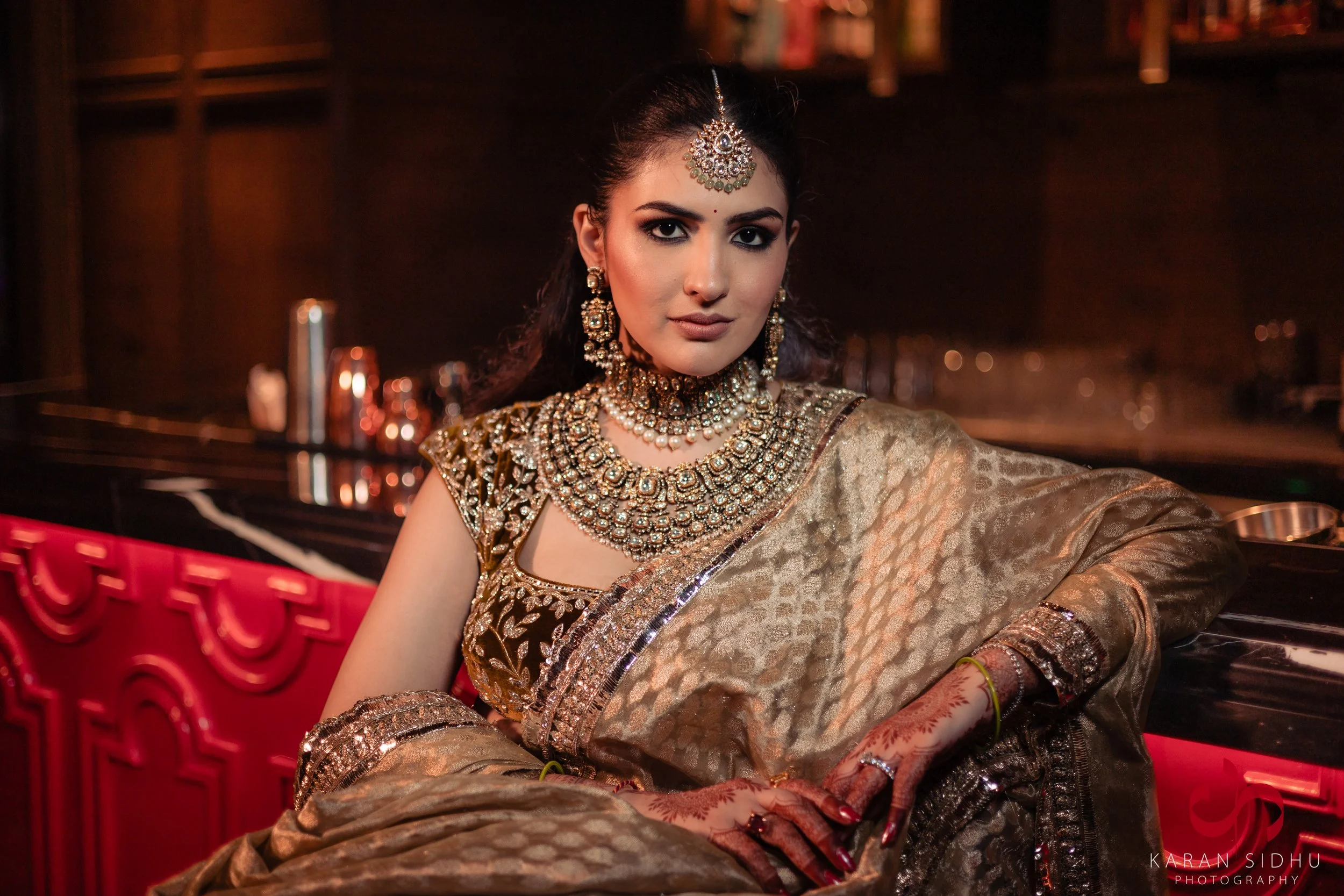 A woman dressed in traditional Indian bridal attire with elaborate jewelry, including a maang tikka, earrings, necklace, and bangles, sitting in a dimly lit setting with a bar in the background.