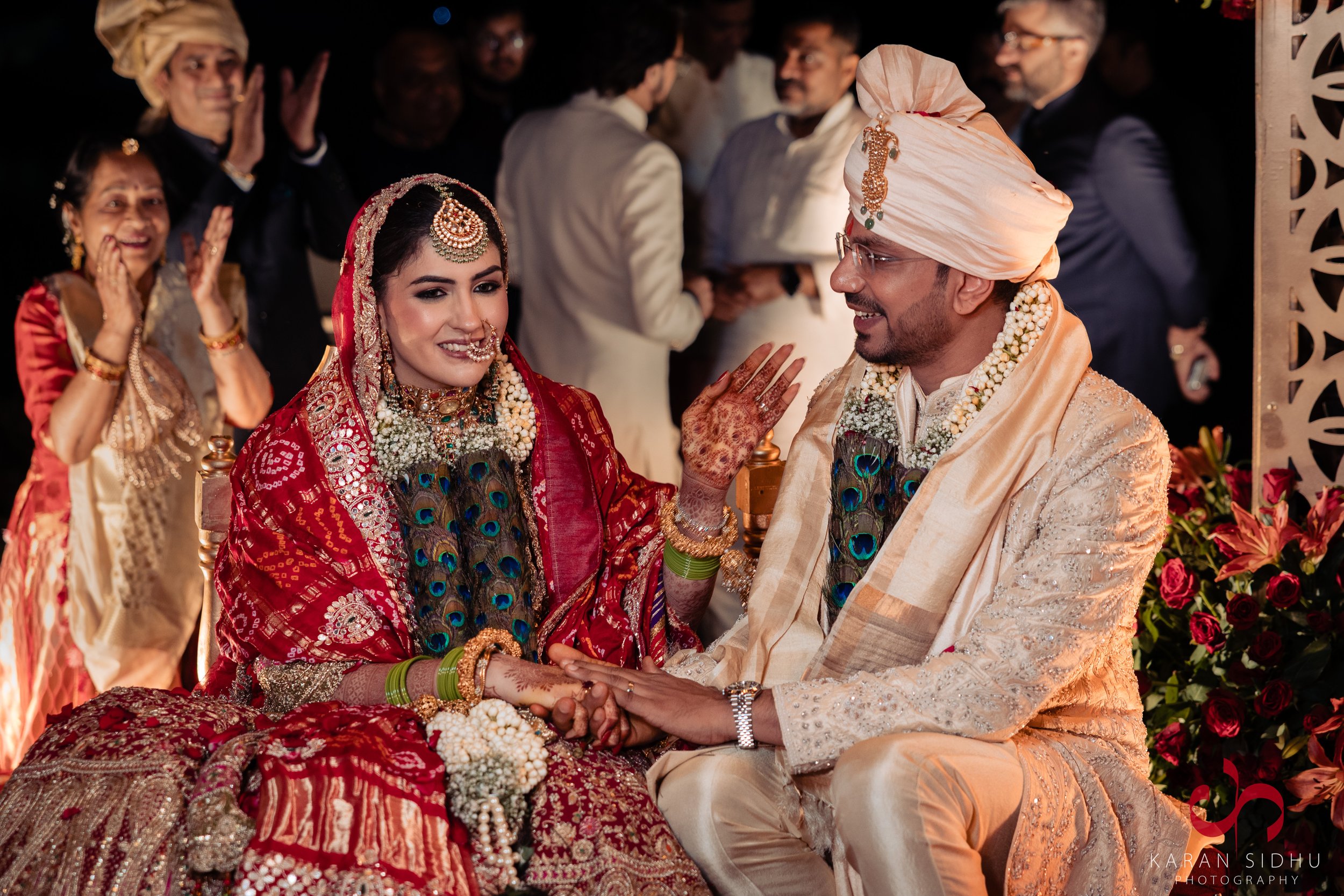 A bride and groom at their traditional Indian wedding ceremony, smiling and holding hands, surrounded by friends and family in colorful attire.
