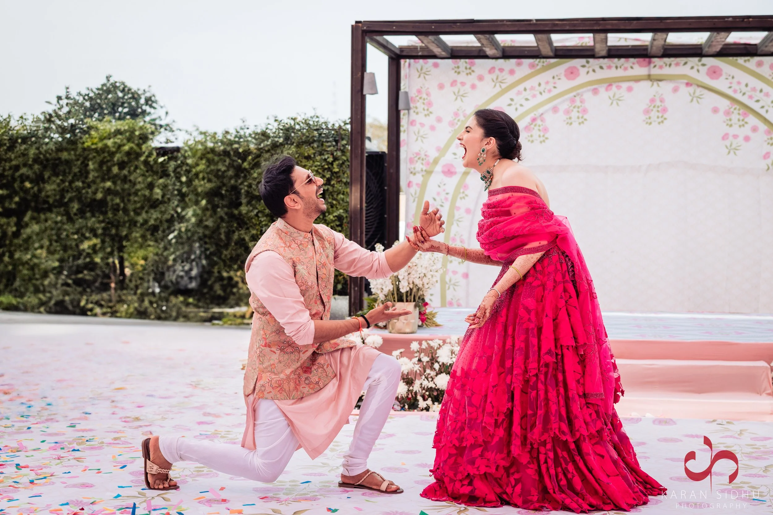 A man proposing to a woman at an outdoor event, with both dressed in traditional Indian attire. The woman is wearing a bright pink lehenga with intricate detailing, and the man is dressed in a light pink kurta with embroidery, kneeling and holding th