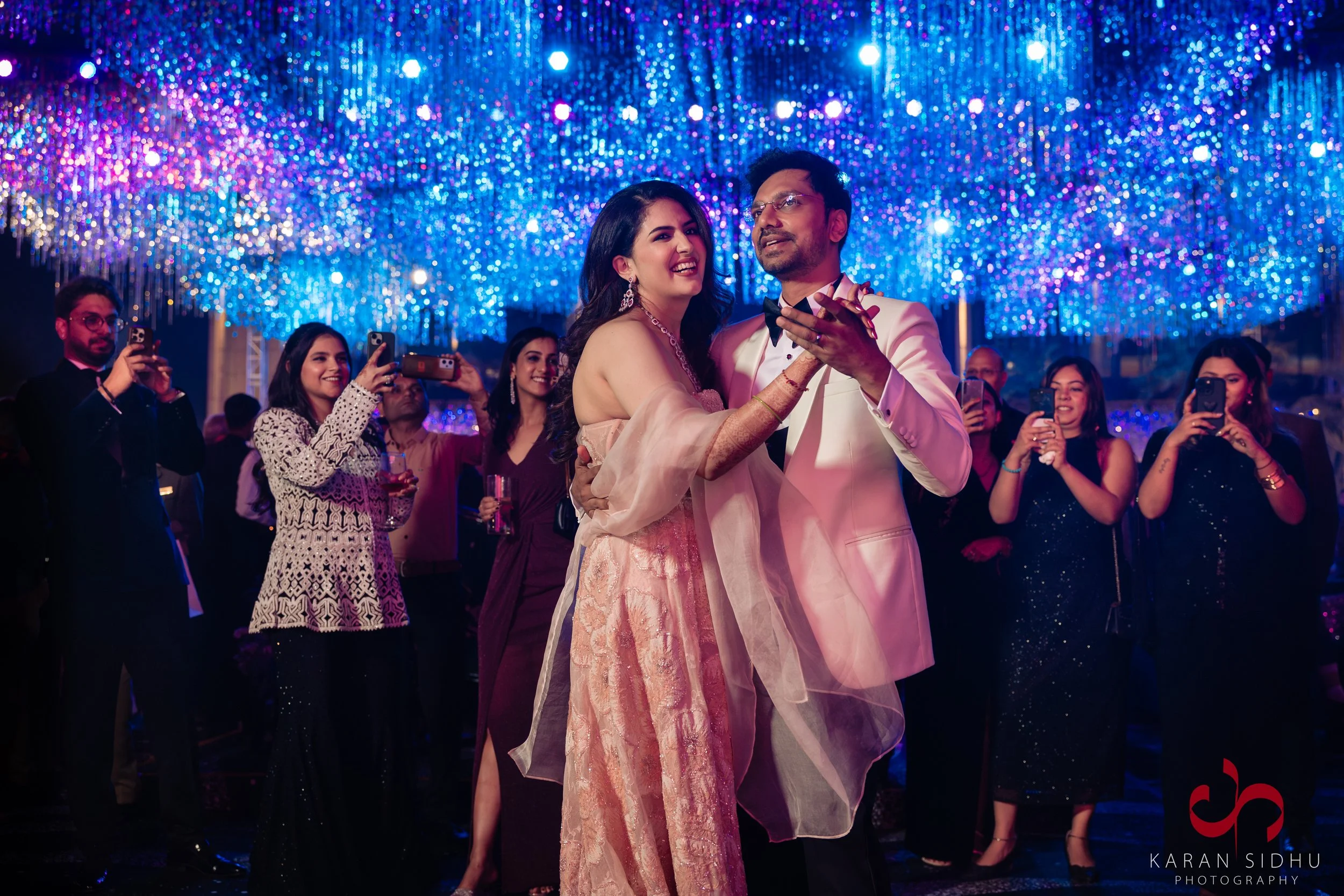 A wedding reception dance featuring a bride and groom dancing under a canopy of colorful lights, with guests capturing the moment on their phones.