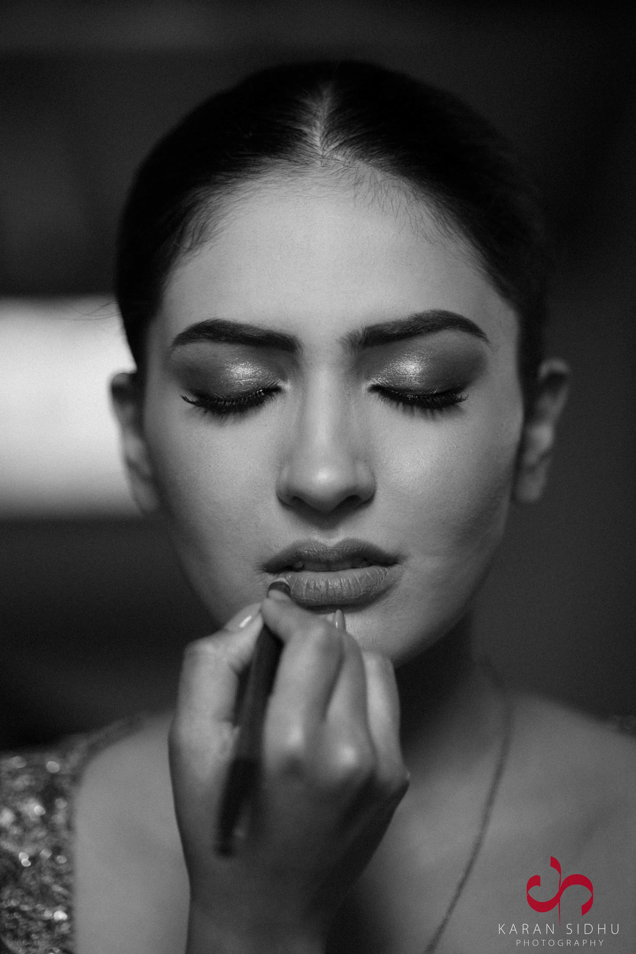 Black and white photo of a woman with closed eyes getting her lips makeup applied by a makeup artist. The woman has dark hair and makeup with shimmering eyeshadow.