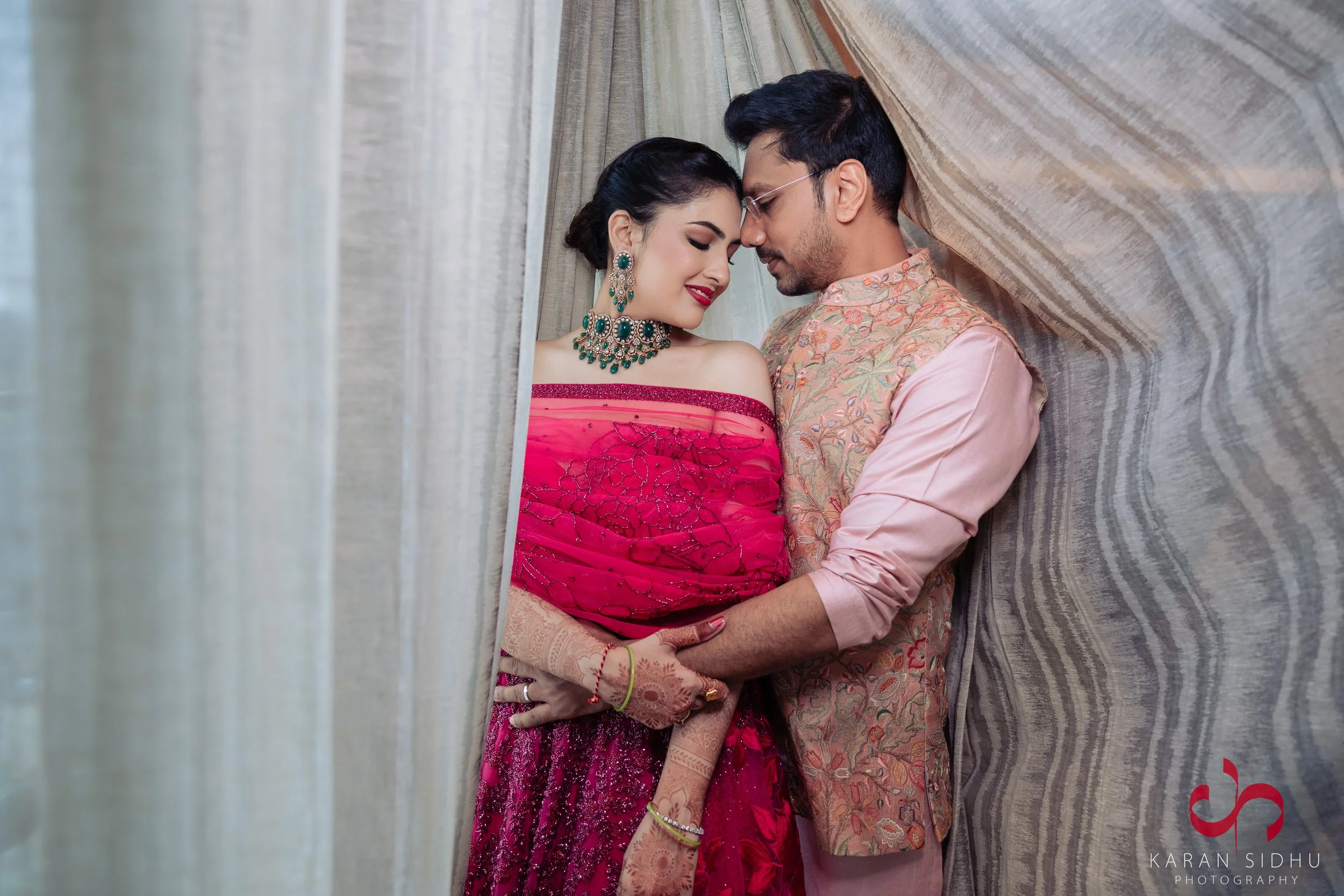 A man and woman dressed in elegant South Asian attire, leaning close together with foreheads touching in an intimate pose, standing behind a curtain with textured wall background.