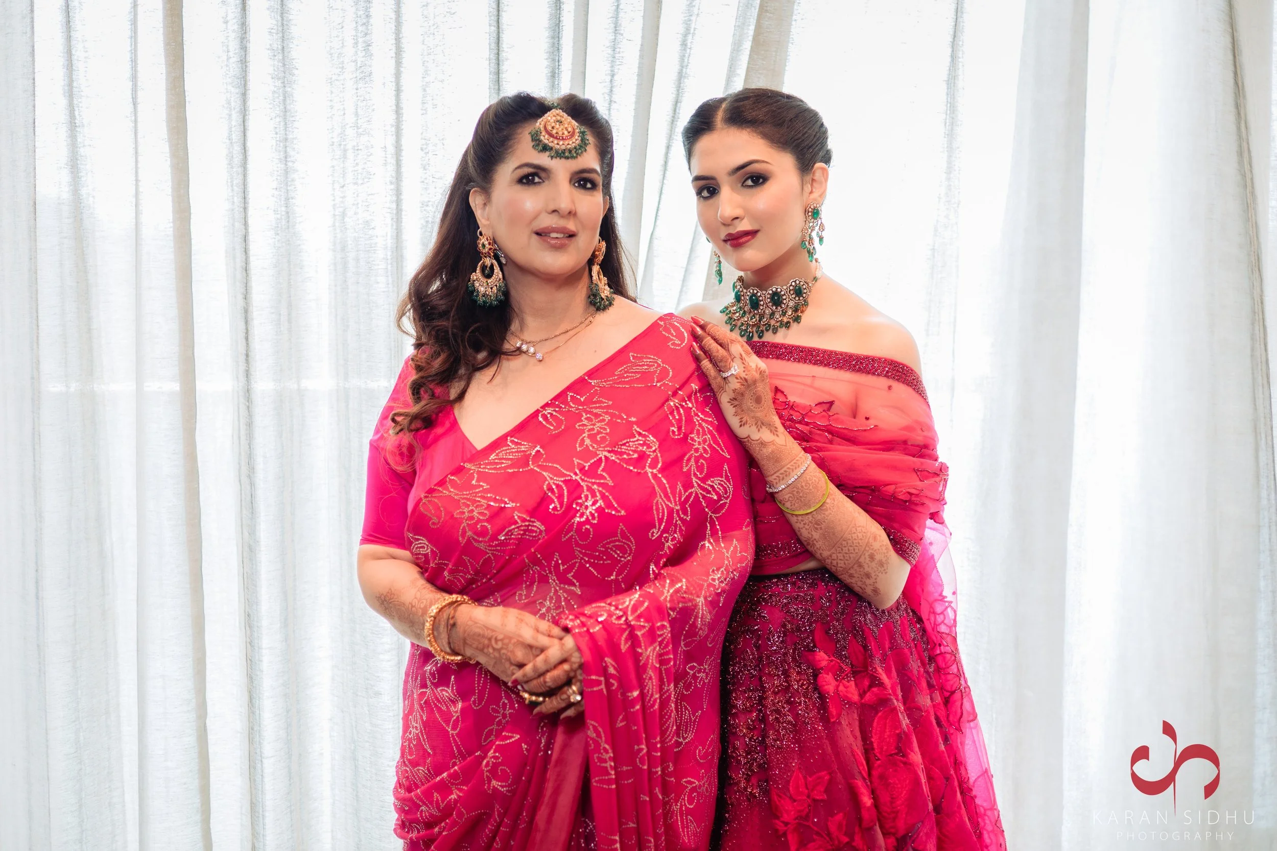 Two women dressed in pink traditional Indian attire and jewelry standing in front of a light-colored curtain.