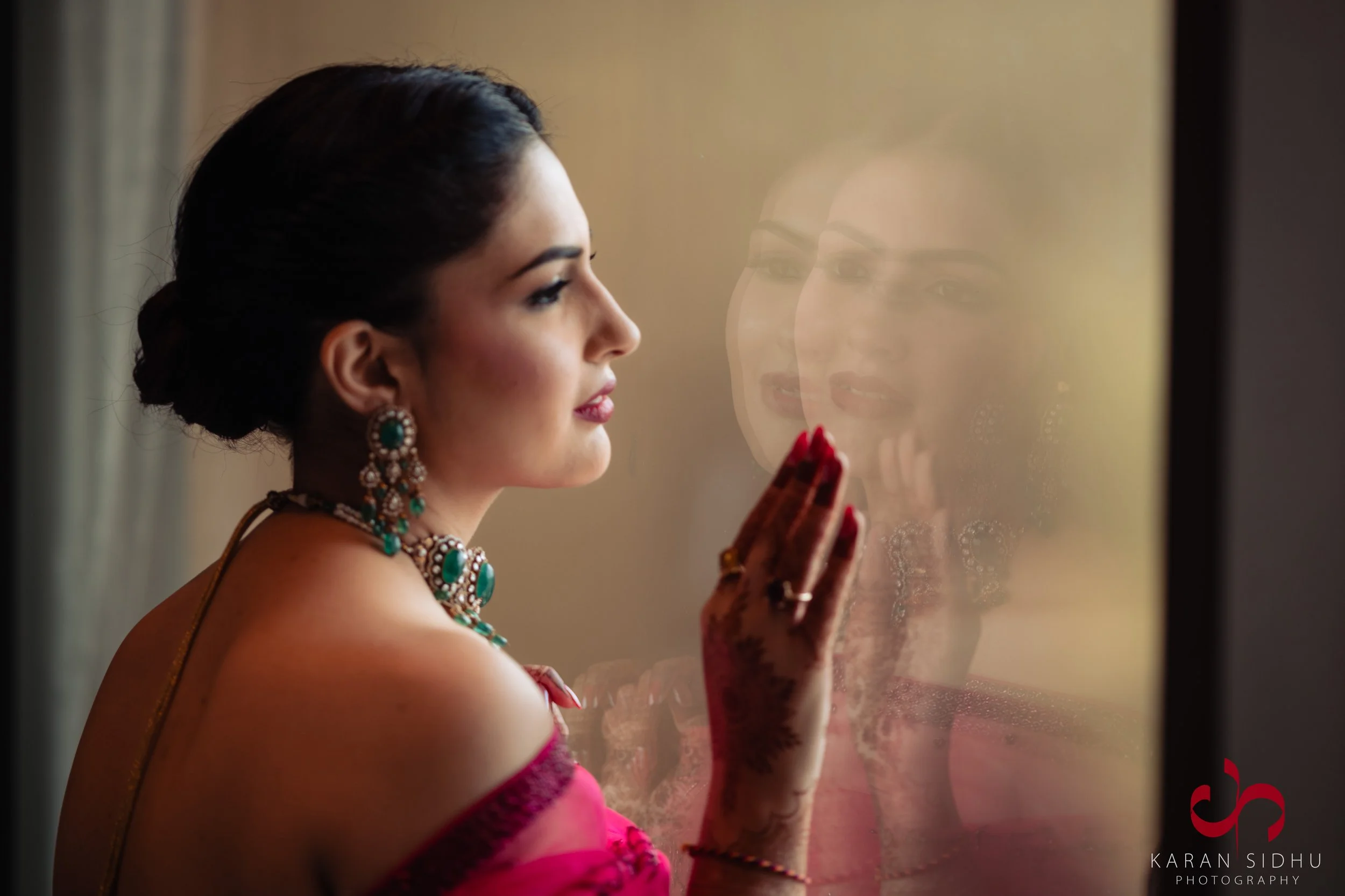A woman dressed in traditional Indian attire and jewelry, standing near a window, looking outside with her hand gently touching the glass, reflecting her face.