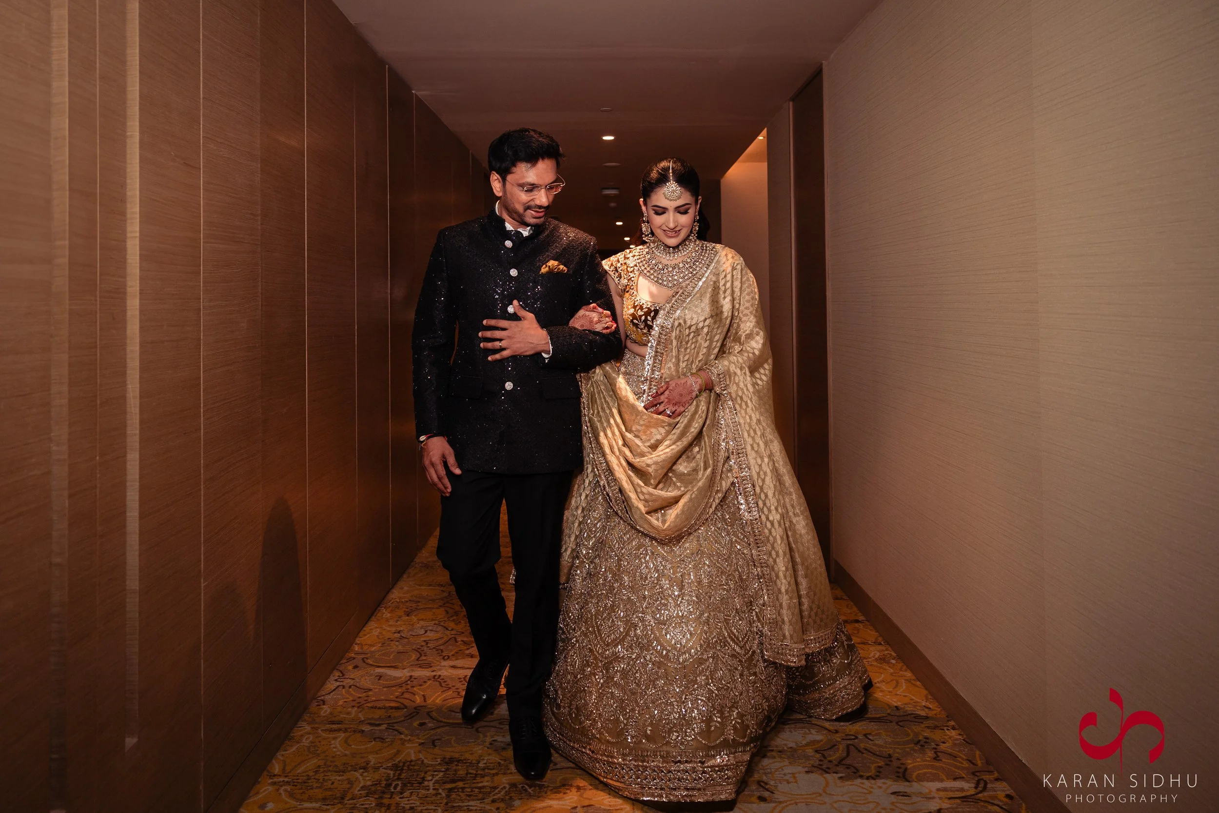 An Indian couple dressed in traditional attire walking down a hotel corridor. The woman wears a gold-colored lehenga with intricate embroidery, and the man wears a black sherwani with embellishments.