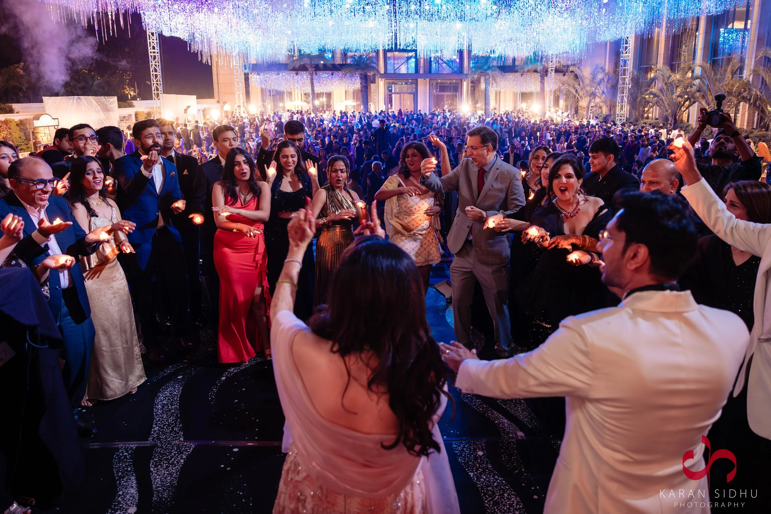 A large gathering of people at a celebration, with some holding candles, dancing, and enjoying at an outdoor event decorated with string lights and hanging decorations, nighttime setting.
