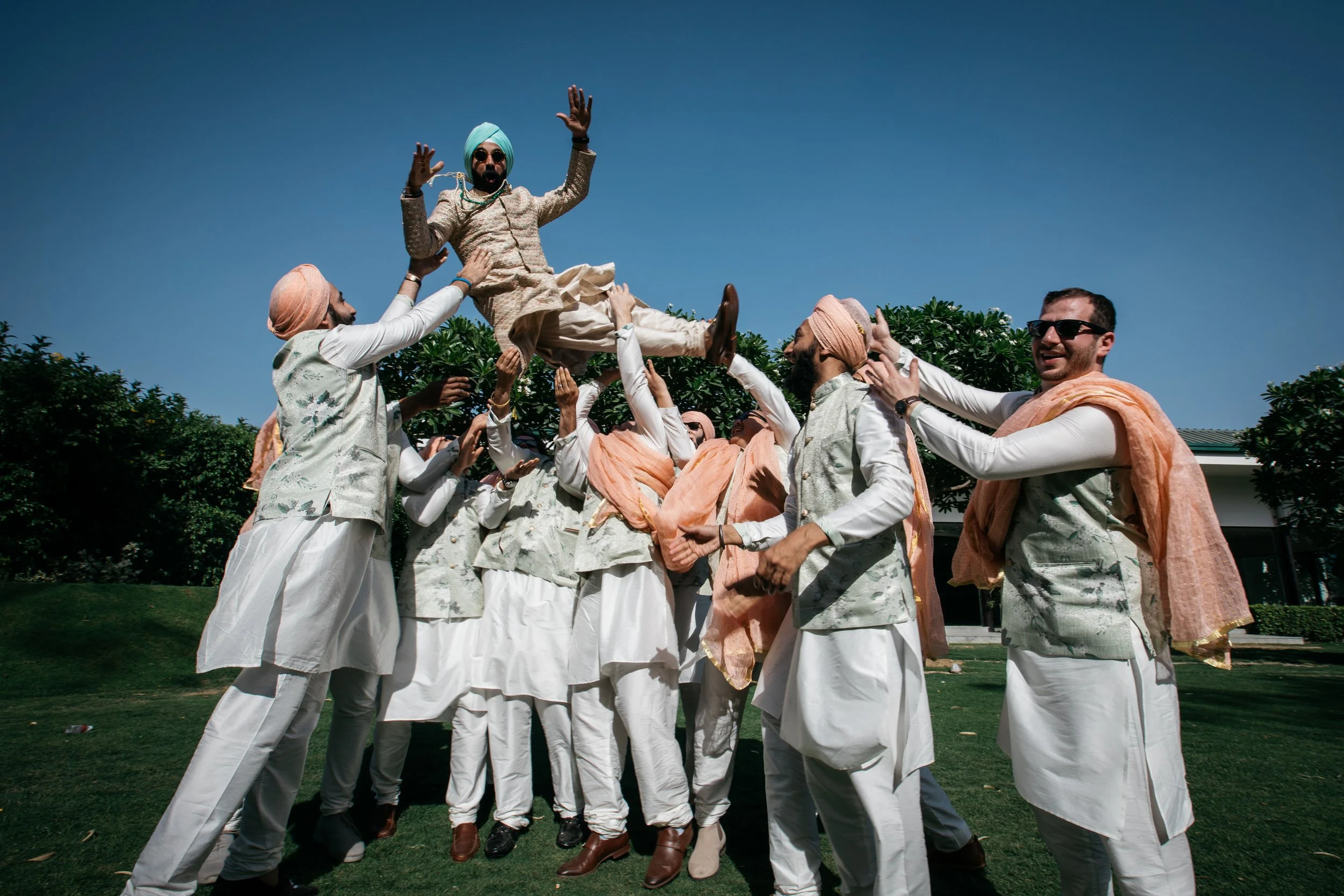 Group of men lifting a man dressed in traditional Indian attire into the air during a celebration outdoors on a clear day.