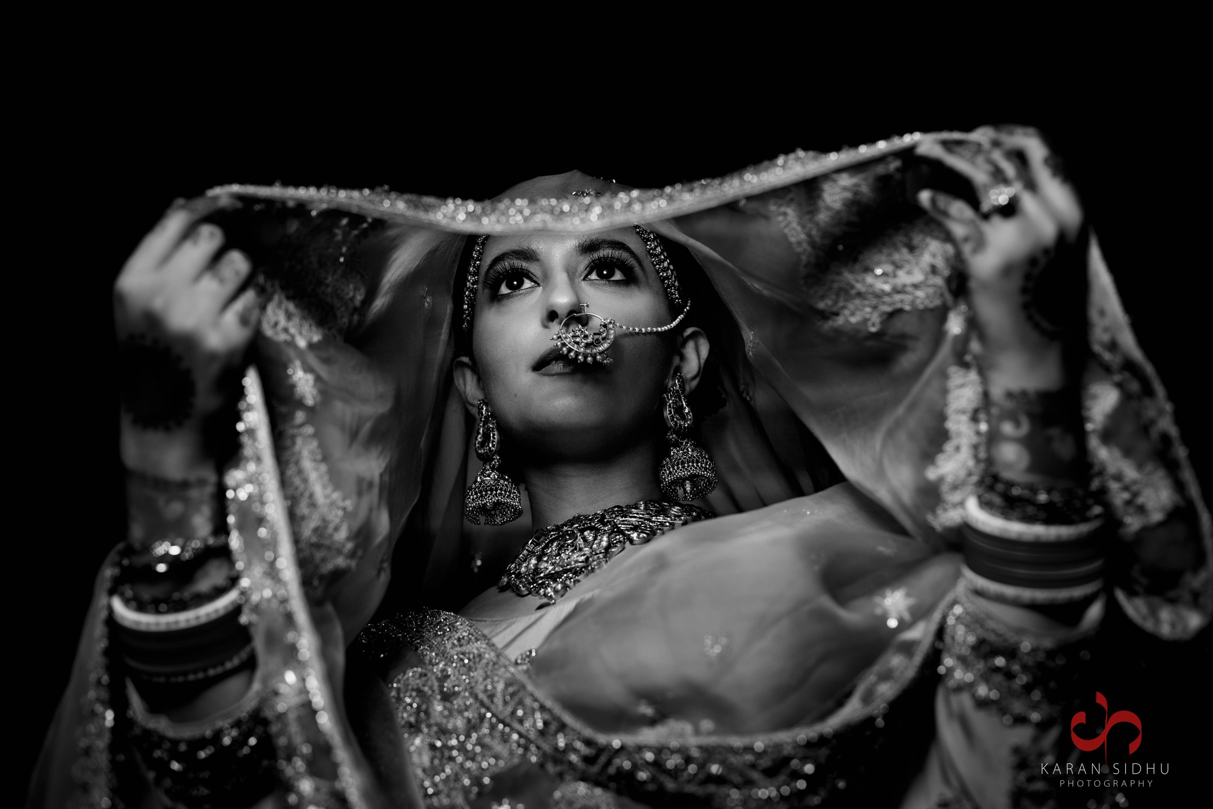 Black and white photo of a woman in traditional Indian bridal attire, holding a sheer dupatta over her head, looking upward with jewelry and makeup visible.