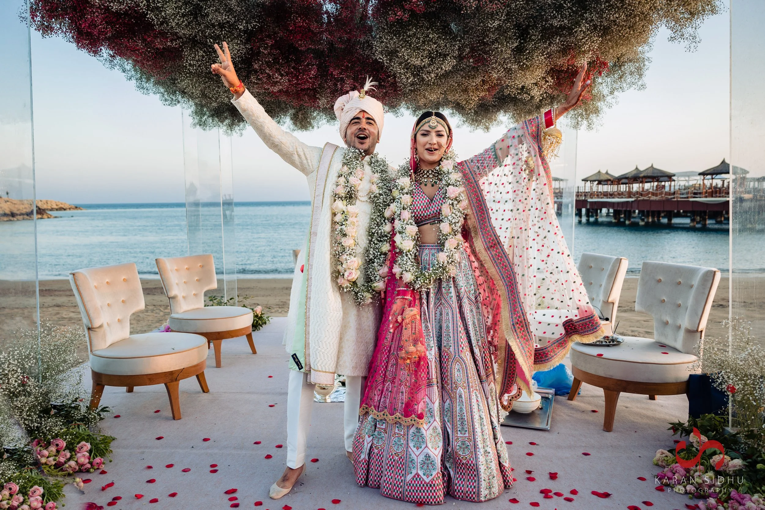 A newly married couple celebrating on a decorated beach wedding stage with the ocean in the background. The bride in traditional colorful Indian attire and jewelry, and the groom in a white outfit with a turban, are smiling and posing with their arms raised.