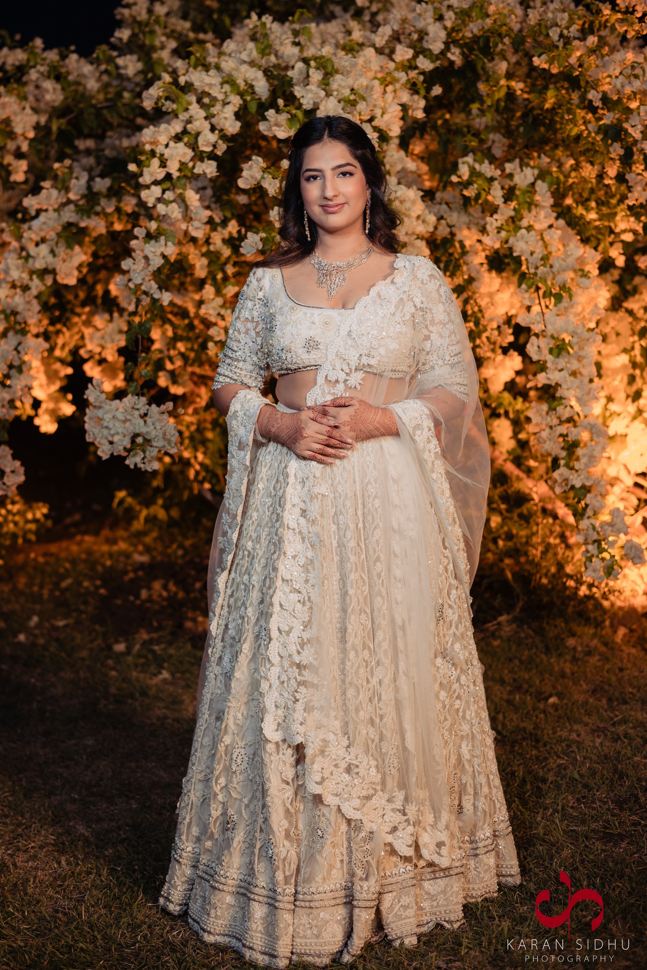 A woman in traditional Indian wedding attire standing in front of illuminated white flowers at night.