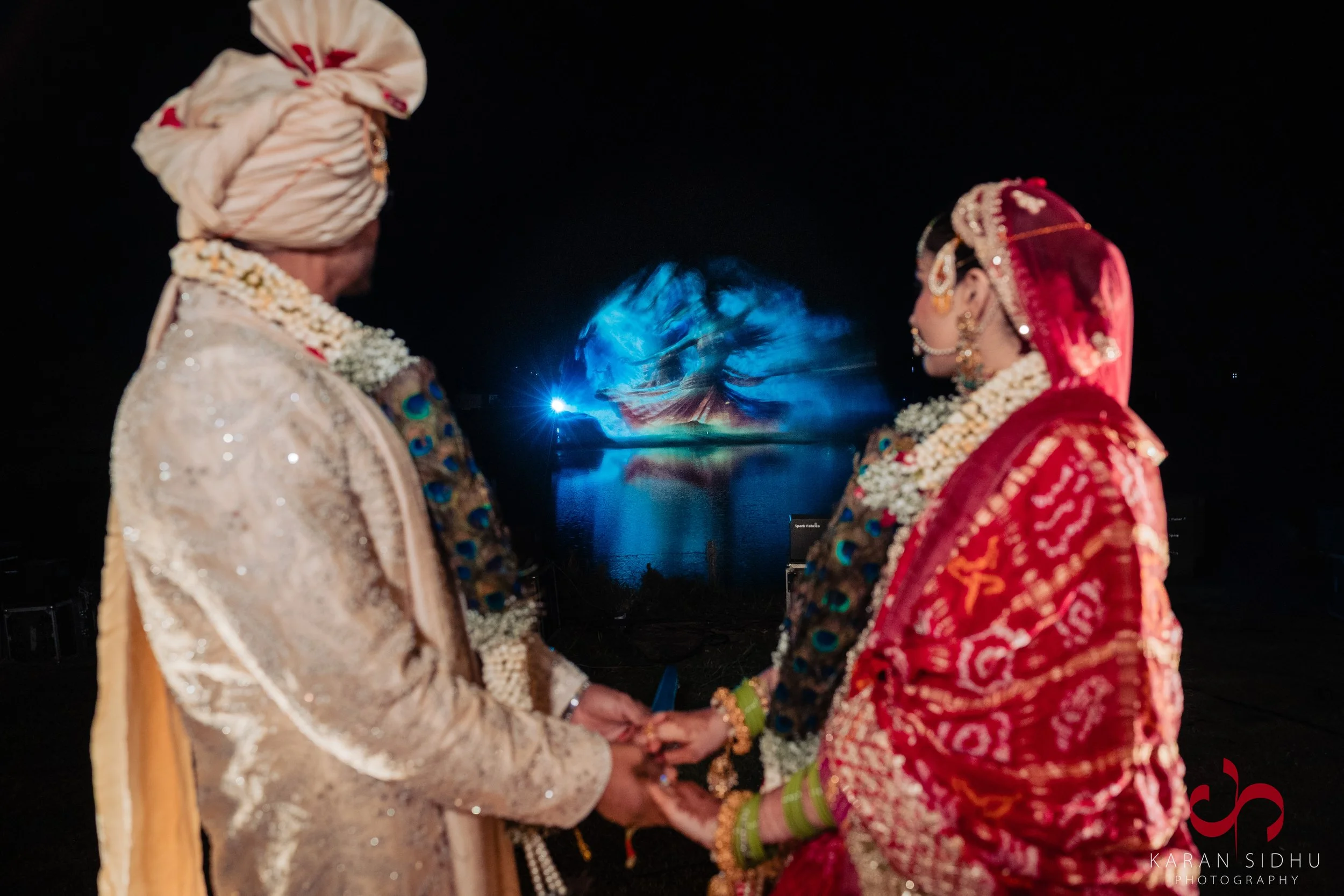 A couple in traditional wedding attire holding hands during a night-time wedding ceremony by a body of water with a lit fountain in the background.
