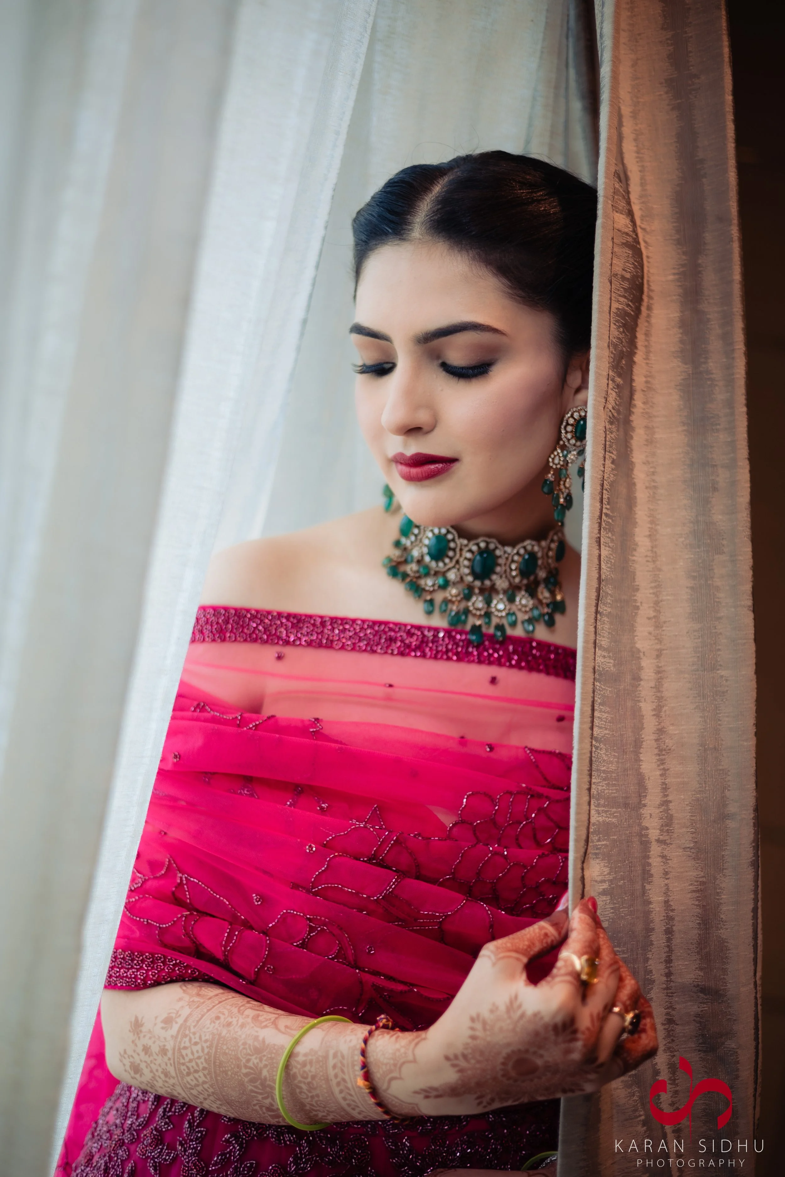 A woman dressed in a vibrant pink saree with intricate embroidery, posing behind a curtain. She is wearing elaborate jewelry, including earrings and a statement necklace, with henna designs on her hands. She is looking down with a calm expression.
