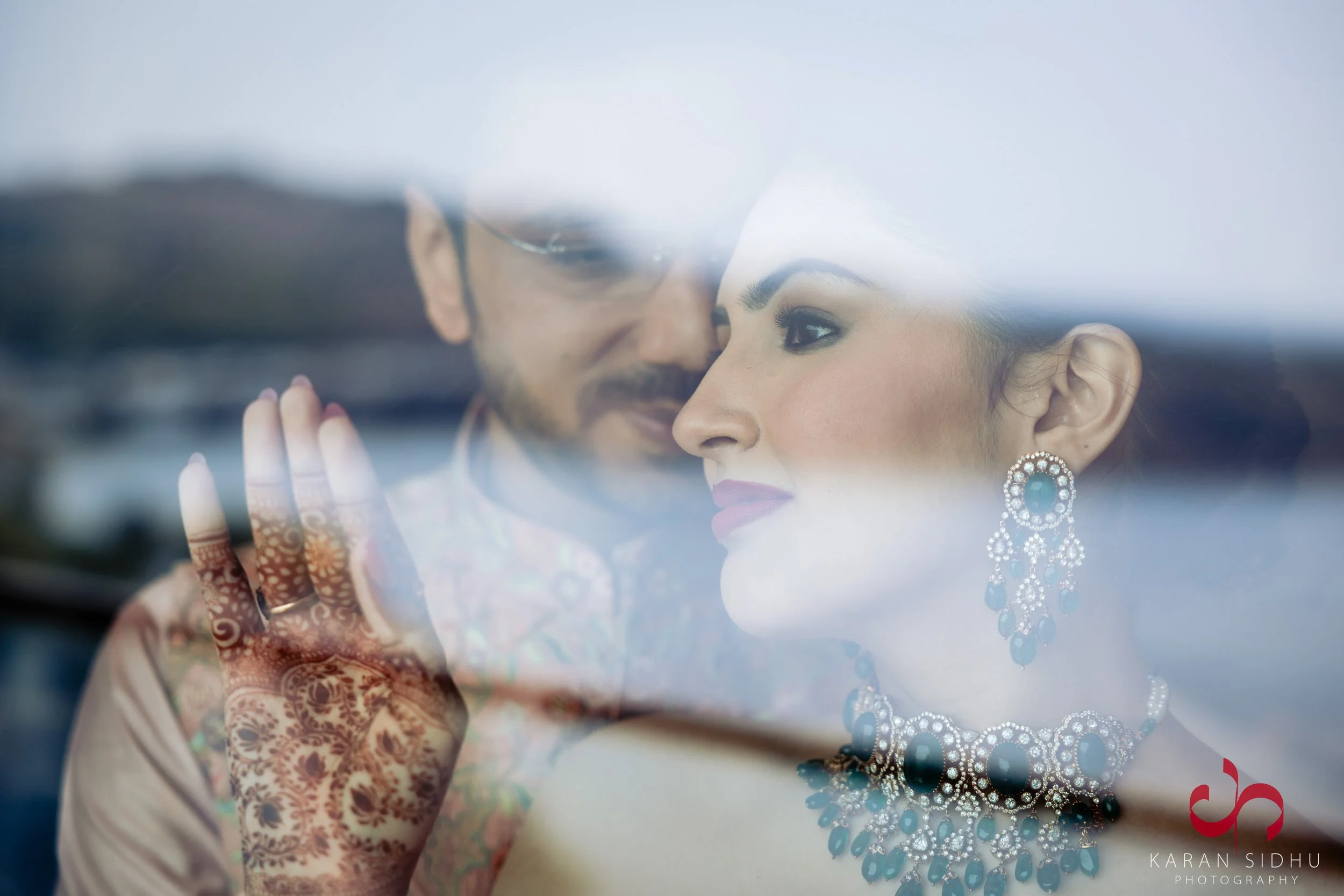 A bride and groom looking at each other through a glass window, with the bride wearing jewelry and the groom touching the glass.