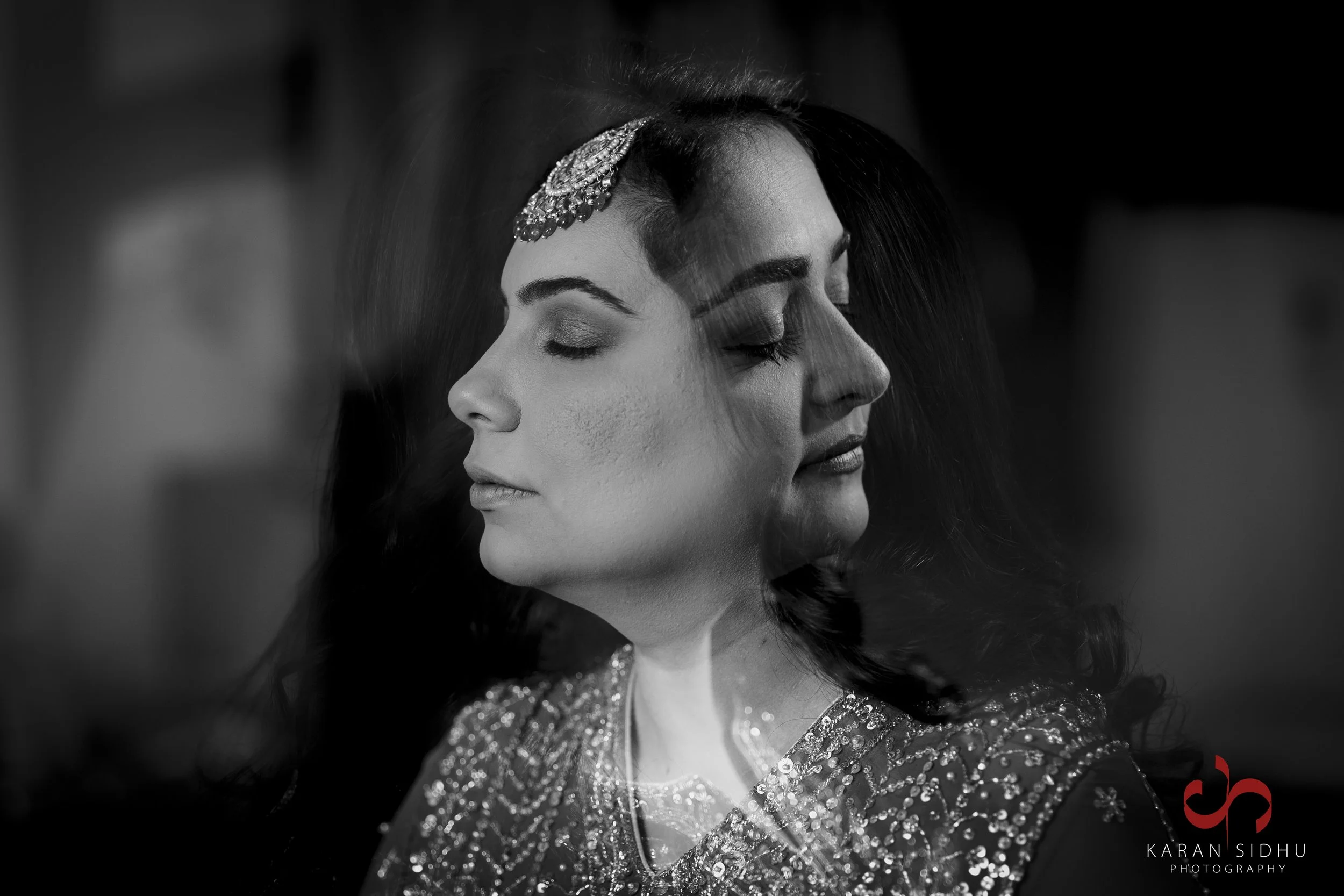 Double exposure black and white photo of a woman with closed eyes, wearing a jeweled headpiece and a decorated outfit, creating an artistic effect.