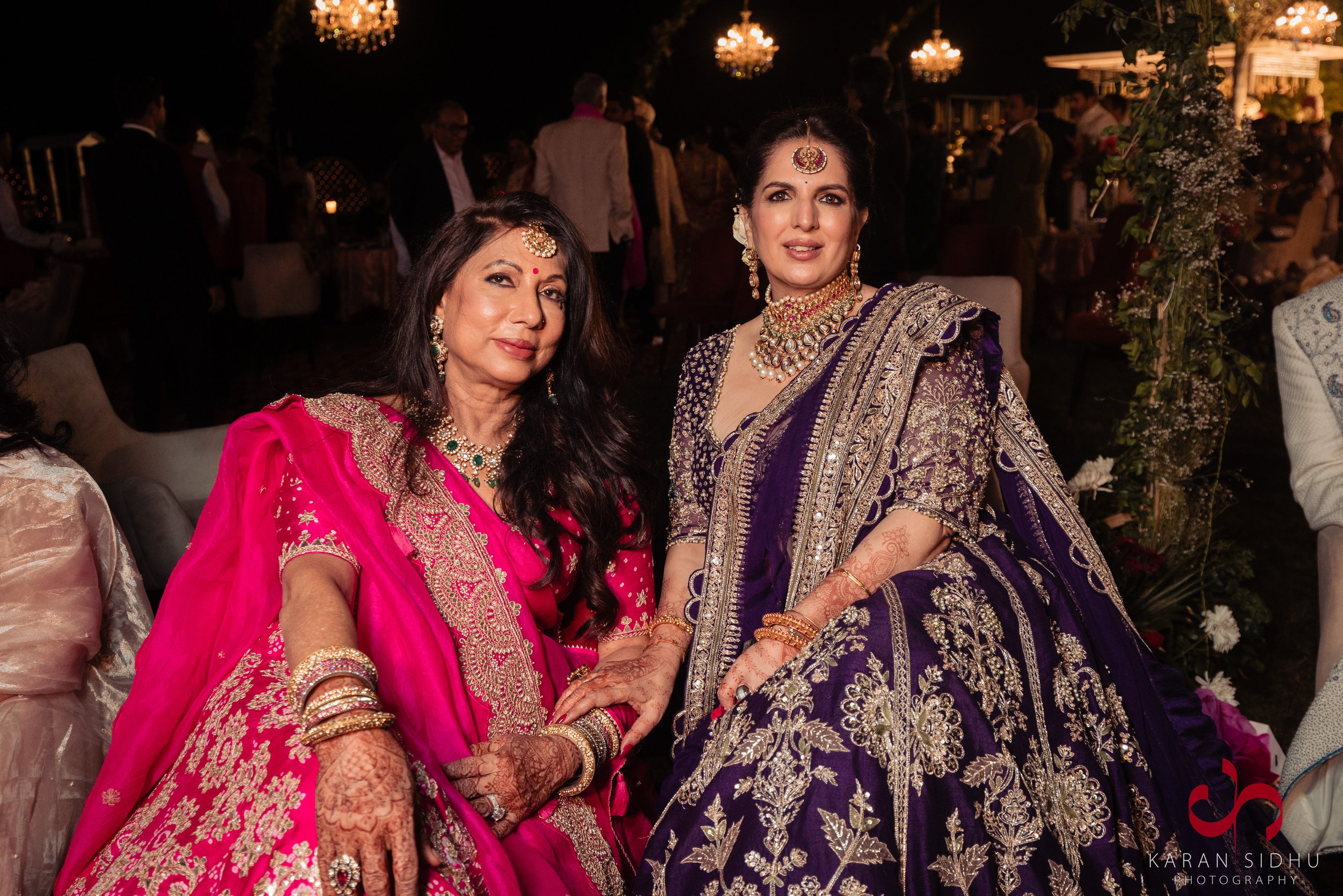 Two women dressed in traditional Indian sarees, adorned with jewelry, attending a celebration at night.