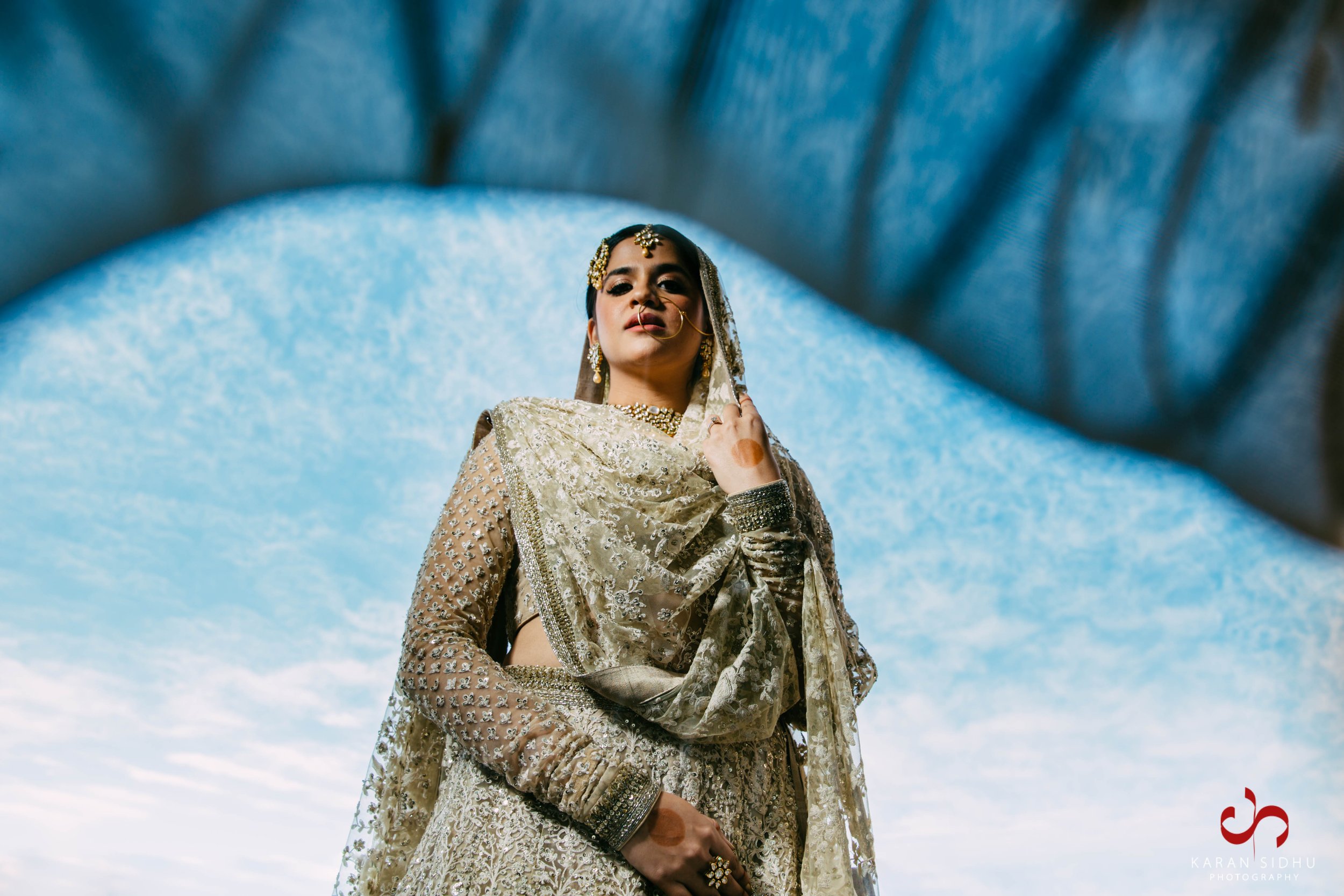 A woman dressed in traditional South Asian wedding attire, with intricate embroidery and jewelry, standing outdoors against a blue sky background.
