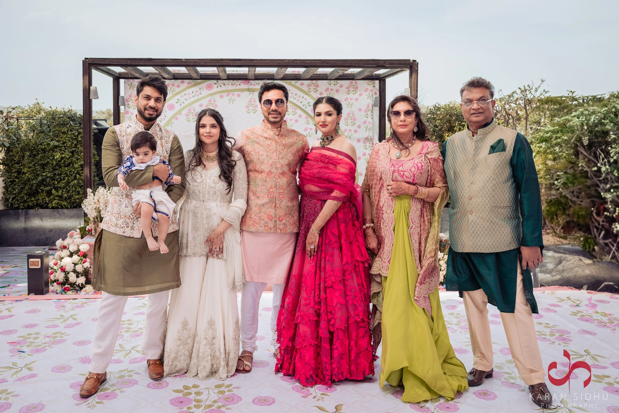 Family of eight, including two young children, standing on a decorated outdoor wedding ceremony stage with floral and greenery backdrop.