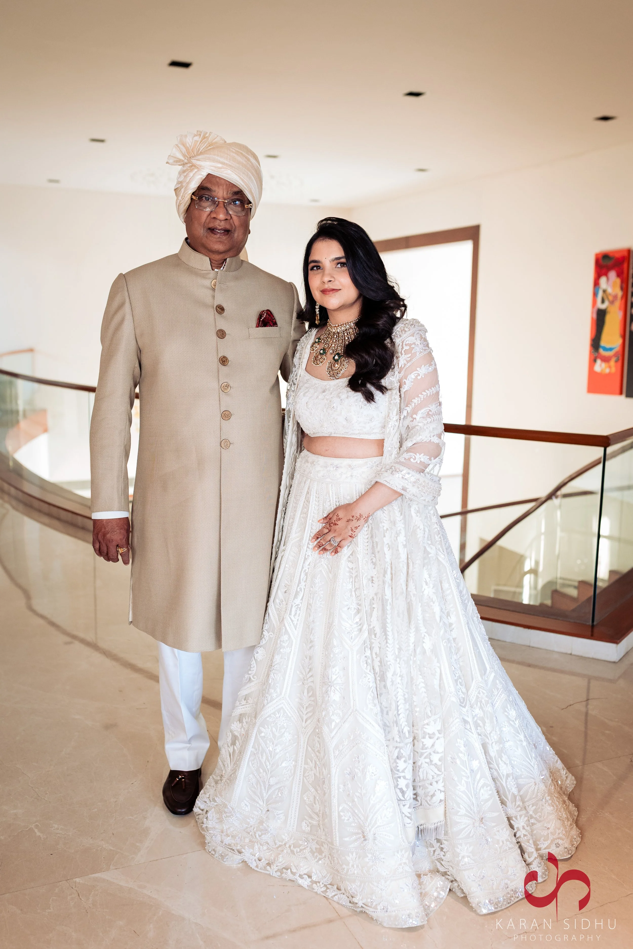 A man in traditional Indian attire and a woman dressed in a white embroidered dress standing together indoors, likely at a celebration or wedding.