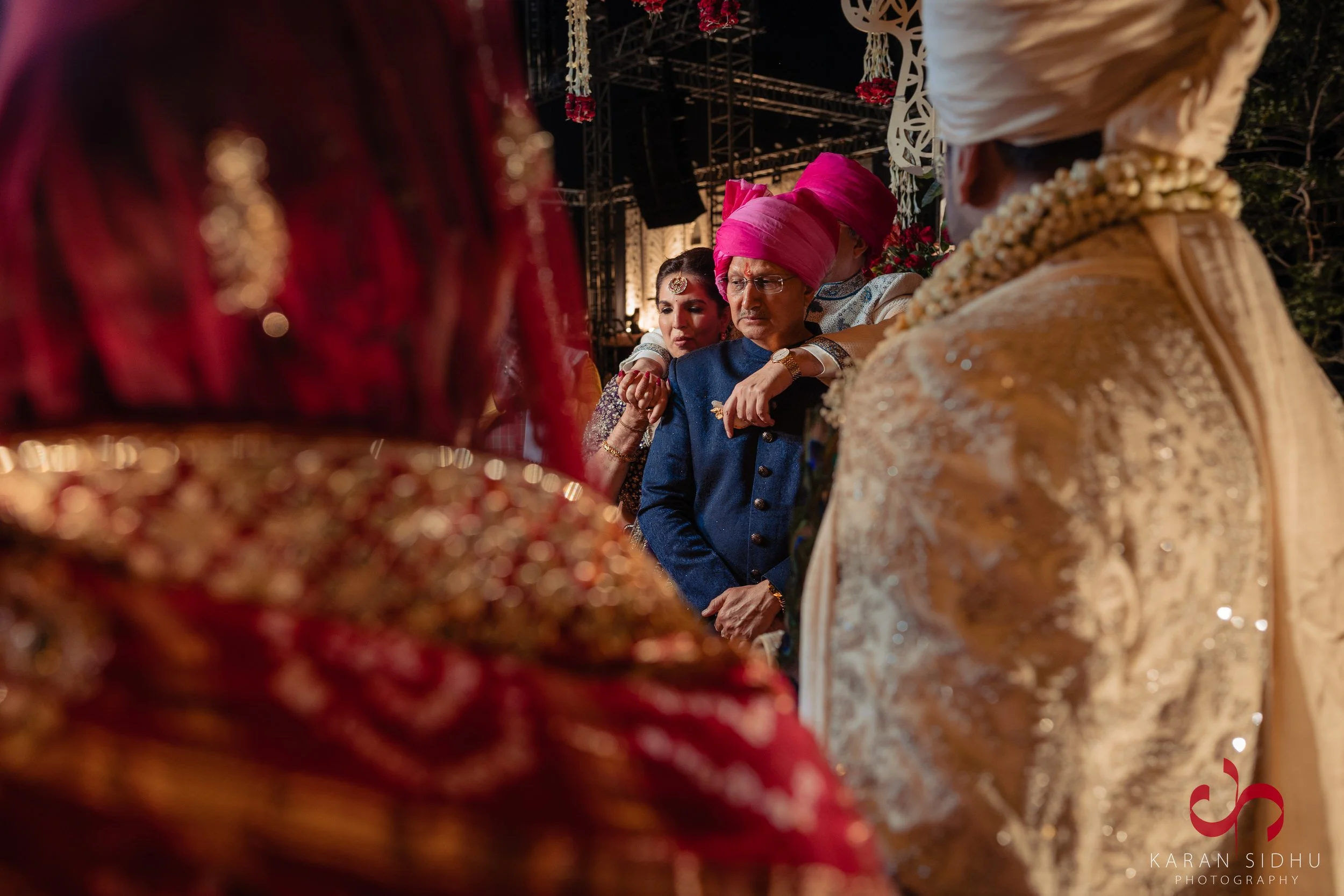 People in traditional Indian wedding attire, standing closely together during a ceremony, with a man wearing a pink turban and a woman with intricate jewelry visible.