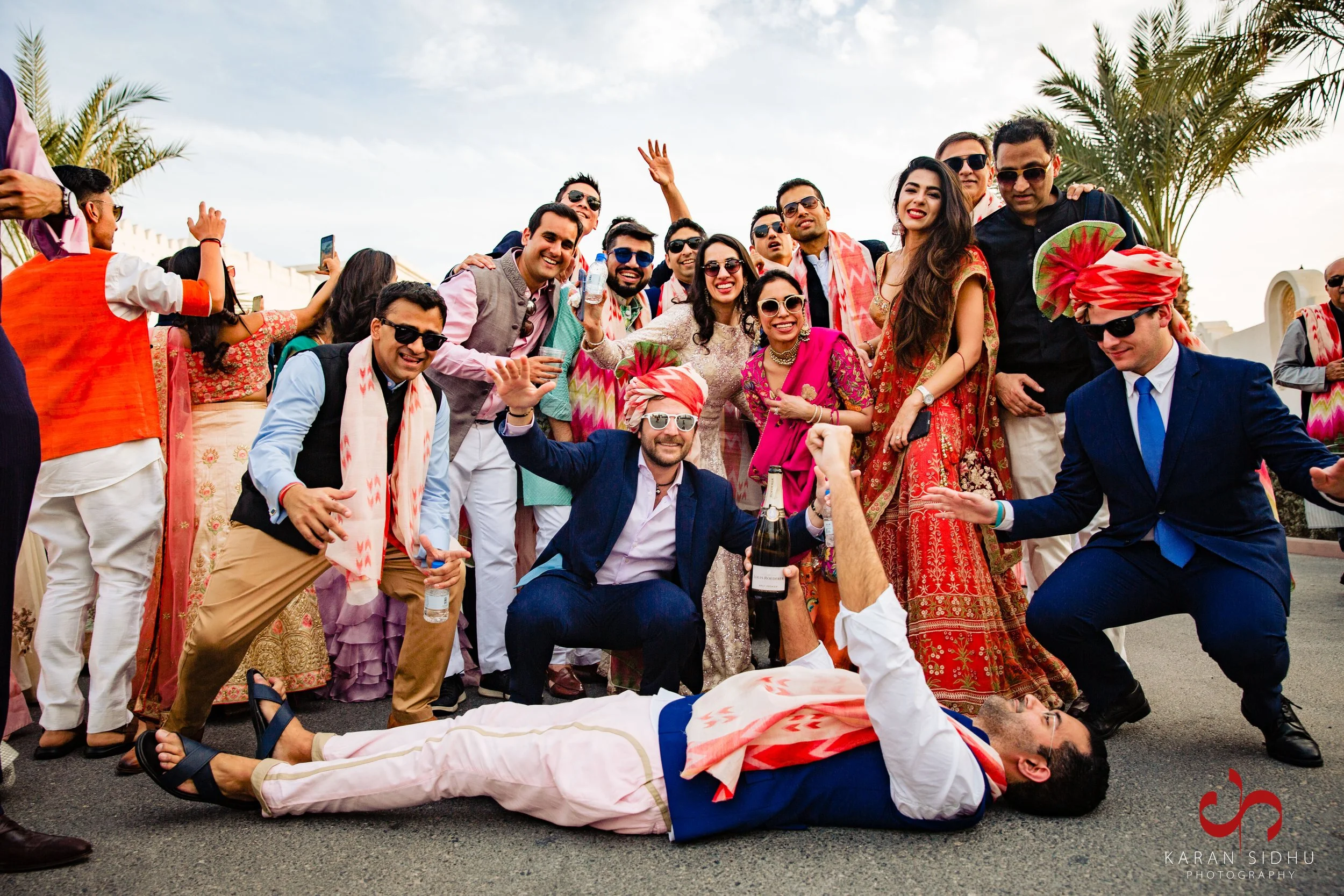A group of people celebrating at a traditional Indian wedding, with some dressed in colorful sarees, sherwanis, and turbans, smiling and posing for a photo outdoors under a partly cloudy sky.