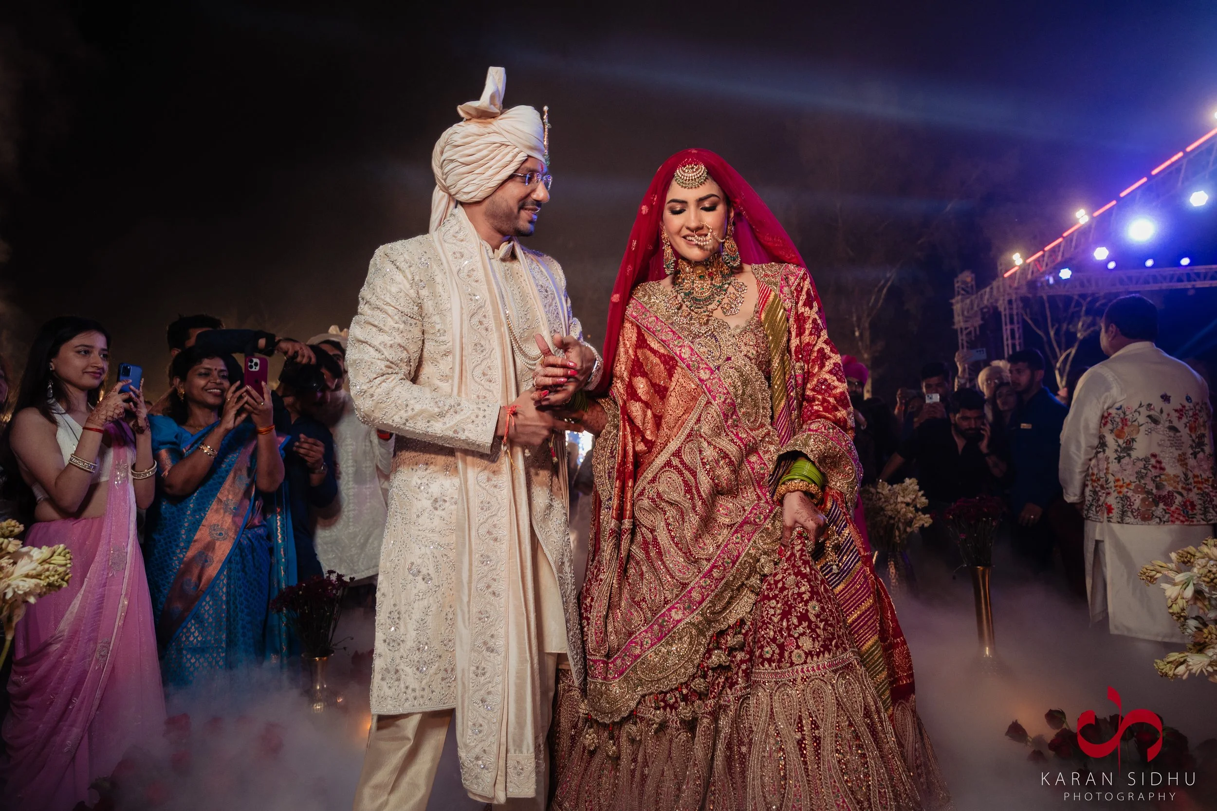Indian bride and groom during wedding ceremony, surrounded by family and friends, with guests taking photos.