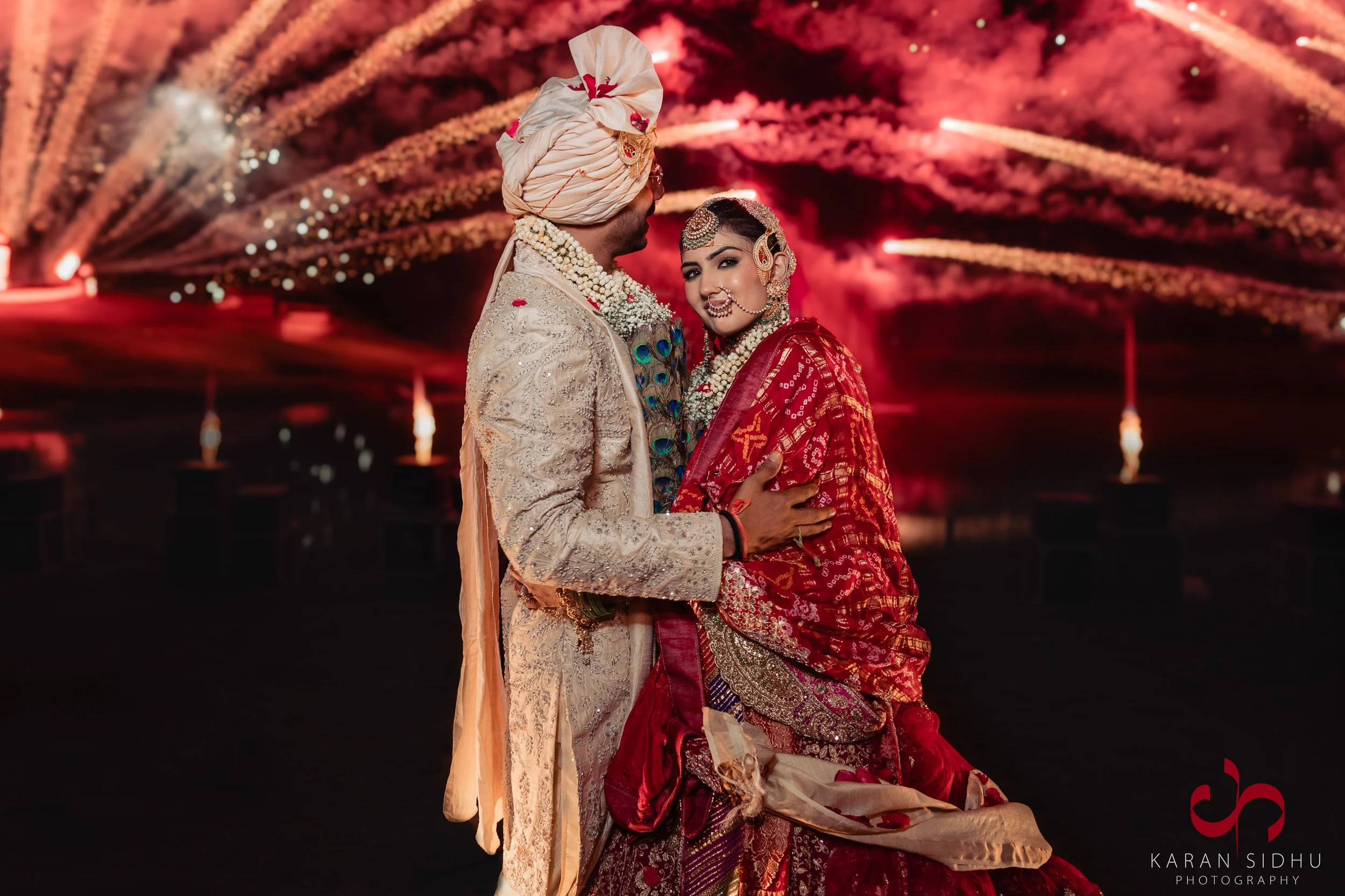 Indian bride and groom dressed in traditional wedding attire celebrating at night with fireworks in the background.