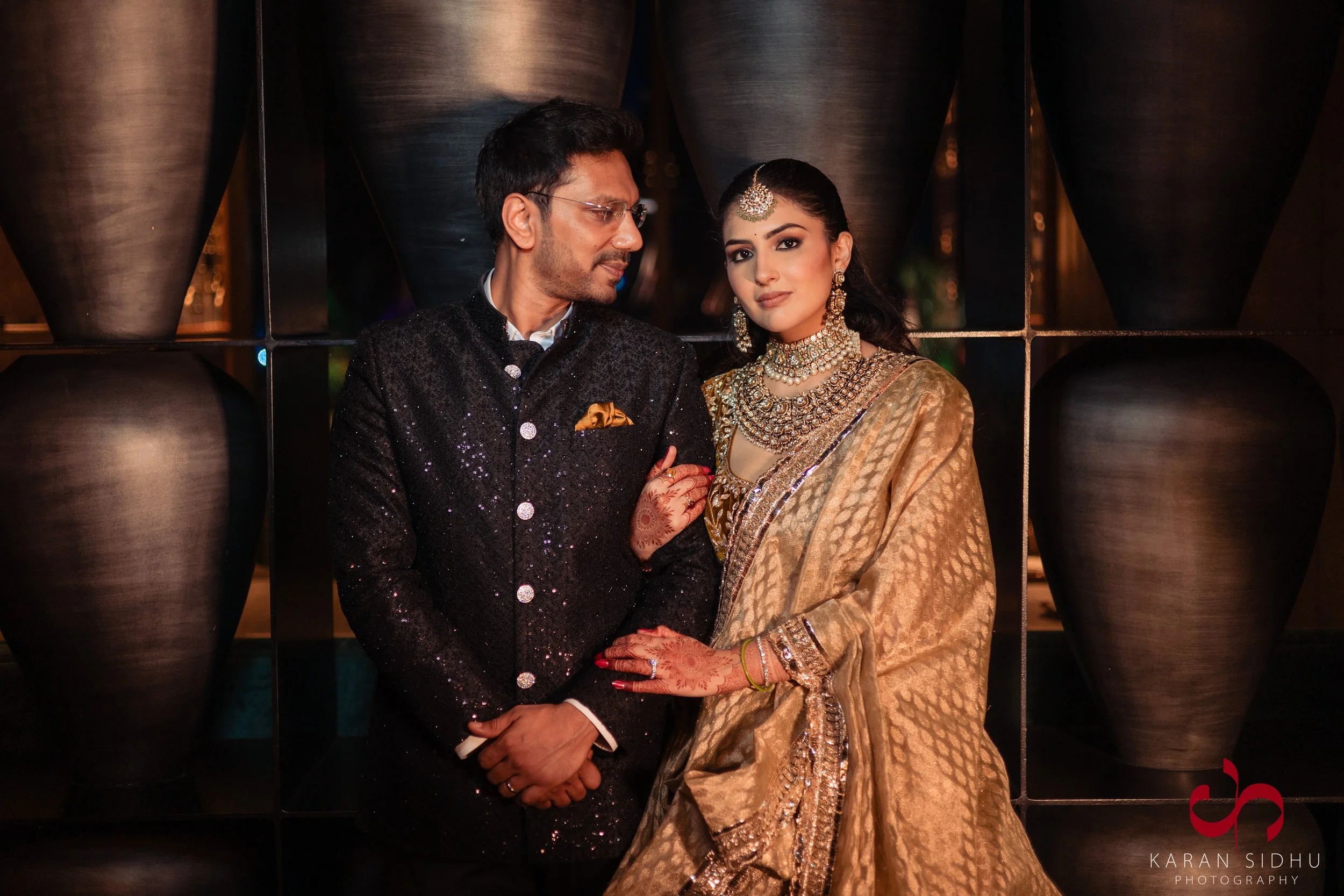 A man and a woman dressed in traditional Indian attire, standing close together indoors with a decorative background of large black pots.