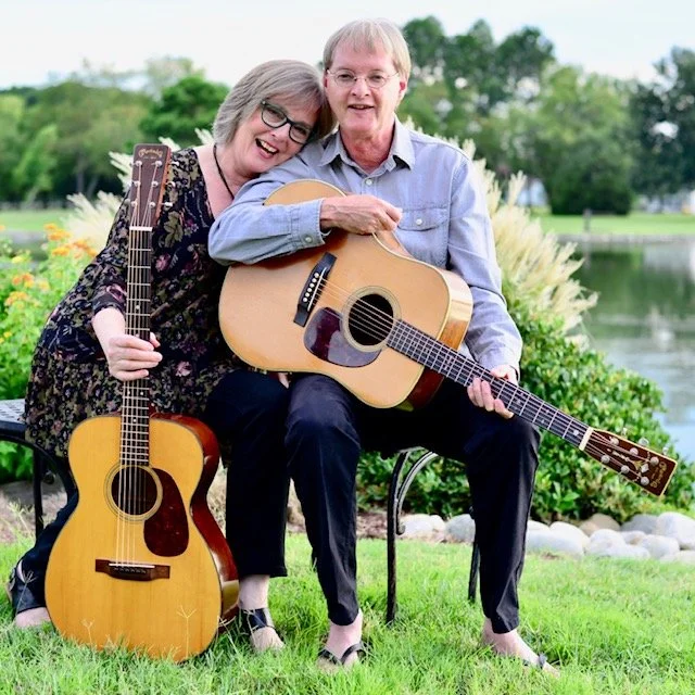 Bill and Pam Gurley, a folk and bluegrass duo from the Northern Neck, sitting together in front of a lake holding acoustic guitars.
