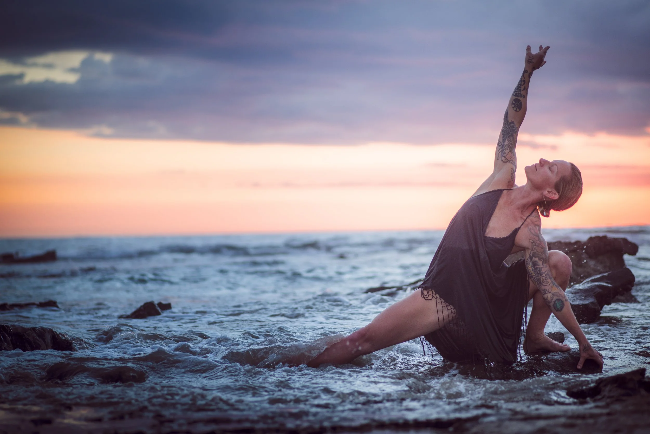Woman with tattoos doing a yoga pose on rocks at the beach during sunset
